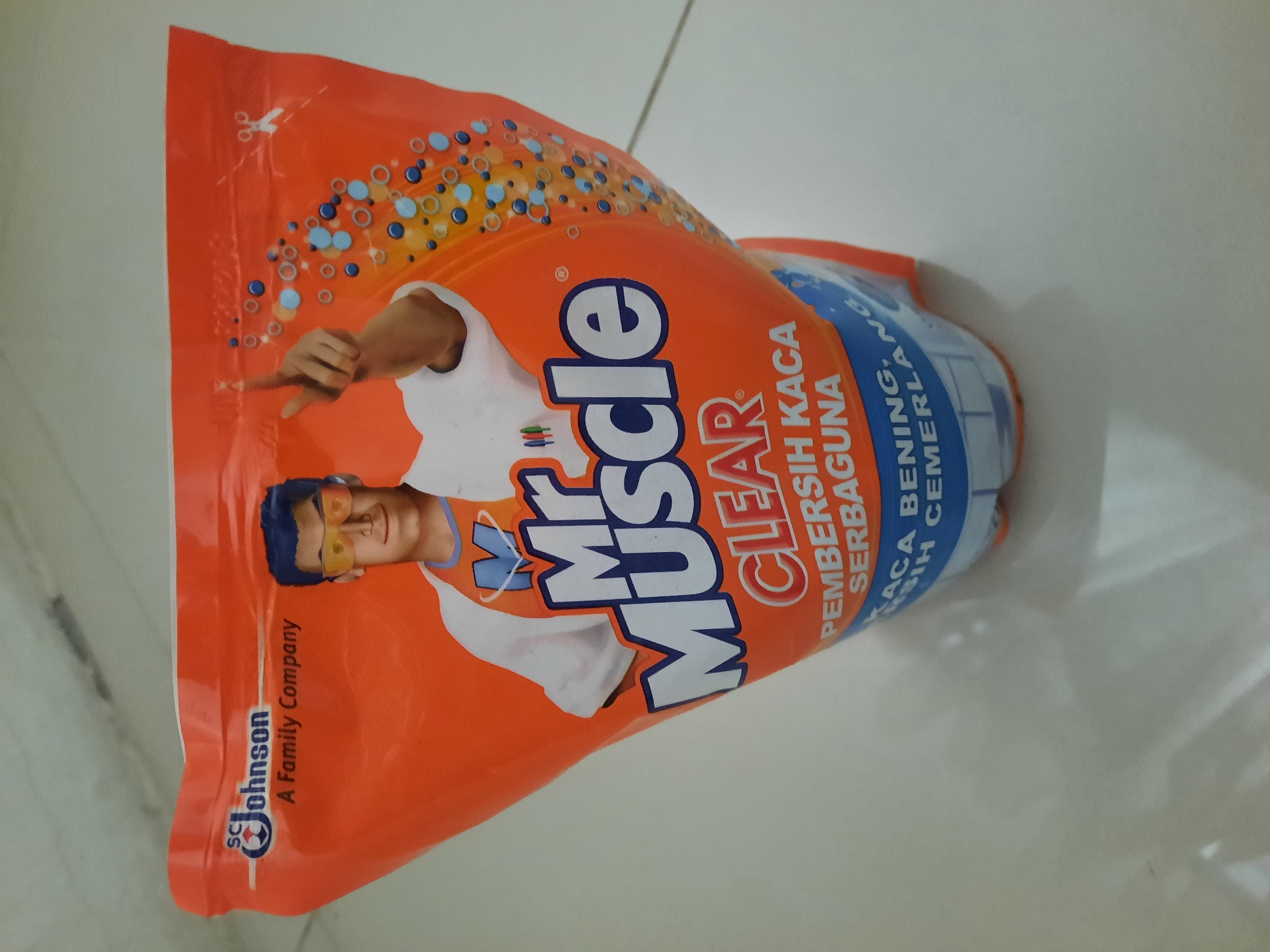 A pouch of Mr. Muscle Clear glass cleaner is displayed upright on a tiled floor. The pouch is predominantly orange with blue and white text and graphics. A cartoon illustration of a man, presumably Mr. Muscle, wearing sunglasses and a lab coat over an orange shirt is featured prominently on the front. He is pointing his right index finger upwards. The text on the pouch reads "SC Johnson A Family Company" at the top left, and "Mr Muscle CLEAR PEMBERSIH KACA SERBAGUNA" in large blue and white lettering. Below this, smaller text reads "KACA BENING, BERSIH CEMERLANG". The background is a light-colored tiled floor. The image does not contain any information about the city of Lhokseumawe, Indonesia.