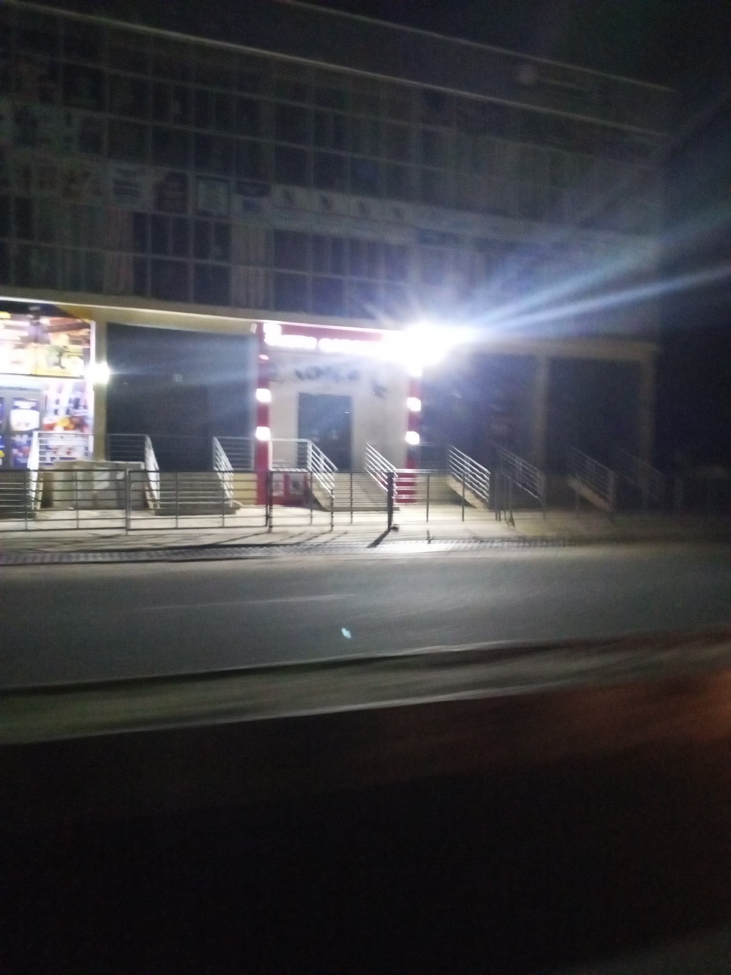 The image depicts a nighttime view of a building exterior in Lokoja, Nigeria. The building appears to be a commercial establishment with multiple levels, featuring a grid-like facade with numerous windows. A bright light source, possibly a streetlamp or exterior lighting, is prominently visible, creating a strong glare and lens flare in the upper right portion of the image. In the foreground, there are several sets of stairs leading up to the building entrance, all equipped with metal railings. A metal fence runs along the sidewalk in front of the stairs. The street in front of the building is paved and appears to be asphalt. The overall scene is dark, with the building and its illuminated entrance being the primary focal points. There are no people visible in the image.
