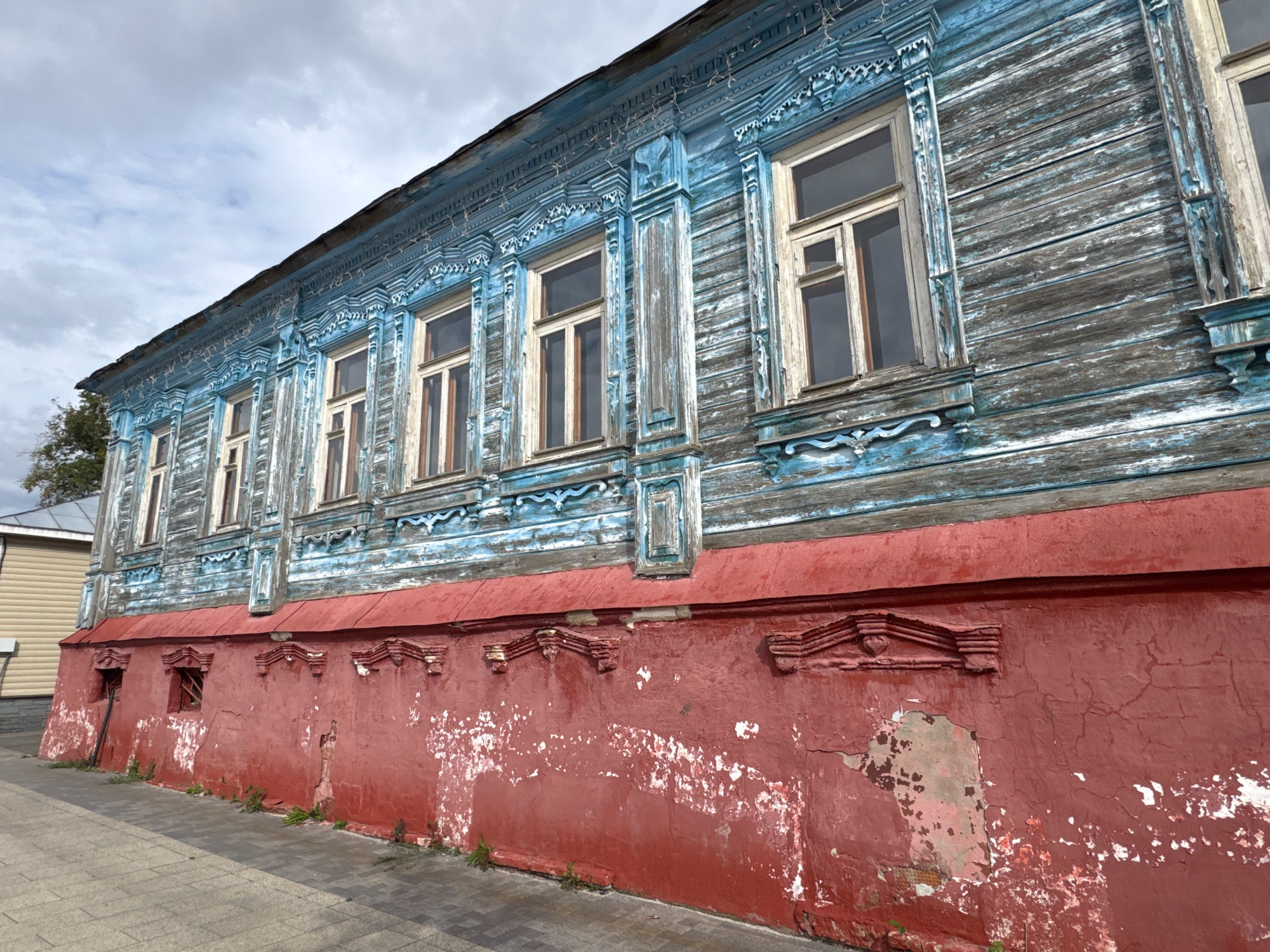This is a side view of an old, two-story wooden house in Zaraysk, Russia. The upper story is painted a faded blue and features ornate wooden trim around the windows and under the eaves. The wood is weathered, with peeling paint revealing grey undertones. The lower story is a rough, red-plastered foundation with several small, barred windows. The plaster is cracked and chipped in places, showing signs of age and disrepair. The house sits on a paved walkway, and a small patch of green weeds grows at the base of the wall. The sky above is overcast with grey clouds. The house appears to be unoccupied and is likely a historic building given its architectural style and condition.