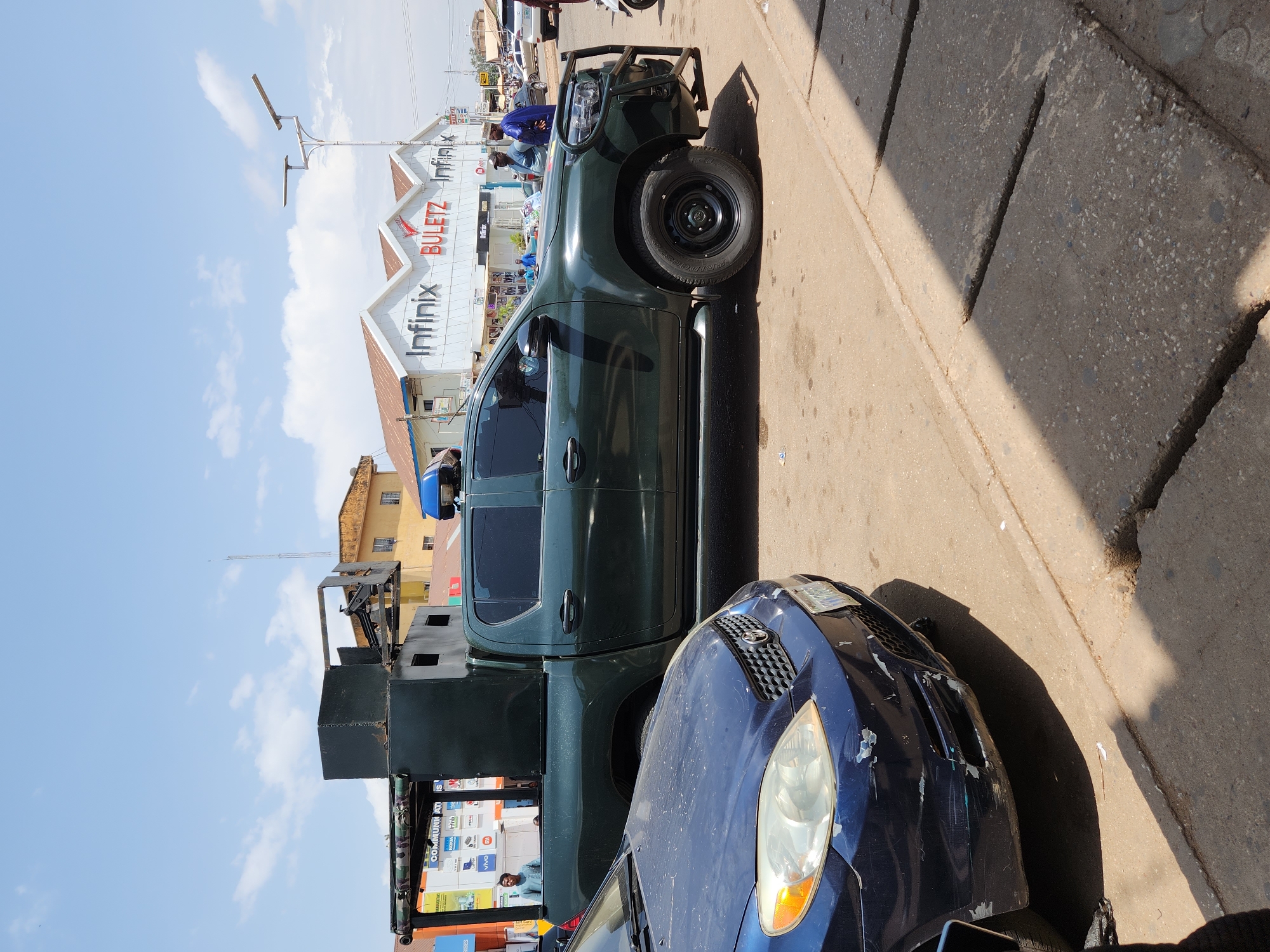 The image depicts a street scene in the city of Unknown, Unknown. A dark green pickup truck with a roll cage and gun turret attached to the truck bed is parked alongside a blue compact car with visible damage to the front bumper and driver's side headlight. The pickup truck appears to be a military or police vehicle. A building in the background has prominent signage displaying "Infinix BULETZ Infinix" in red and white lettering. People are visible in the background, some wearing blue clothing, near the building. The sky is blue with scattered white clouds. The road and curb are visible in the foreground. Shadows are cast on the road surface.