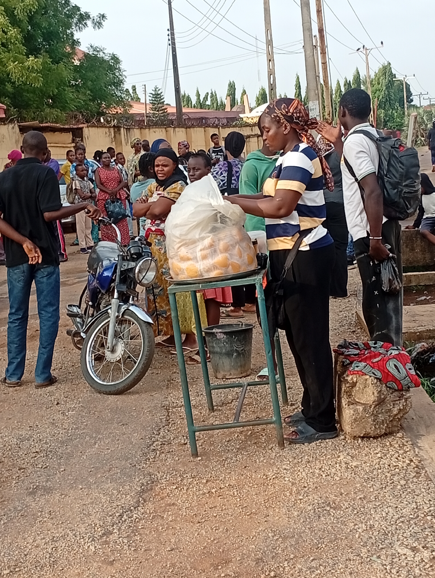 In Barden Yana, Nigeria, a crowd gathers on a gravel road. A woman in a striped shirt and headscarf stands behind a green metal cart laden with a plastic bag full of what appears to be fried dough or bread. She seems to be serving customers. To her right, a man with a backpack waits. To her left, a man in a black shirt and jeans stands beside a parked motorcycle. A bucket is on the ground next to the cart. A stone or concrete block, draped with patterned fabric, is at the edge of the road. In the background, more people are visible, along with a building and trees. Utility poles rise into the sky.