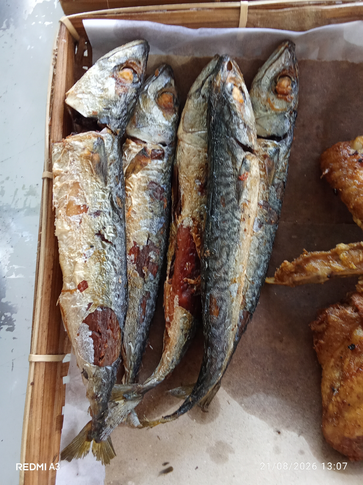 A close-up, overhead shot captures a basket of fried fish arranged on brown paper. The basket, made of woven bamboo, holds at least five whole fried fish, their scales shimmering and their bodies appearing dry and slightly browned. Several pieces of fried chicken, golden brown and appearing crispy, are visible to the right of the fish. The background is a mottled gray surface. A timestamp in the lower right corner reads "21/08/2026 13:07," and "REDMI A3" is in the lower left. The location is specified as Banaran, Indonesia.