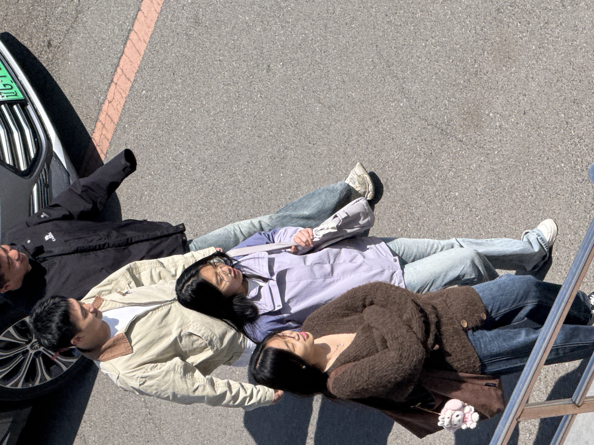 Three individuals lie head-to-toe on an asphalt surface. The first person, a male, is dressed in a black jacket and tan coat, with his head resting on the front tire of a black and silver car. The second person, a female, is wearing a purple shirt and jeans. The third person, a female, is wearing a brown sweater and jeans, with a brown bag and a stuffed animal beside her. A portion of a metal ladder is visible on the right side of the frame. A section of an orange line marking is visible in the top of the frame. A car's license plate is visible on the left. The location is in Zhongtun, China.