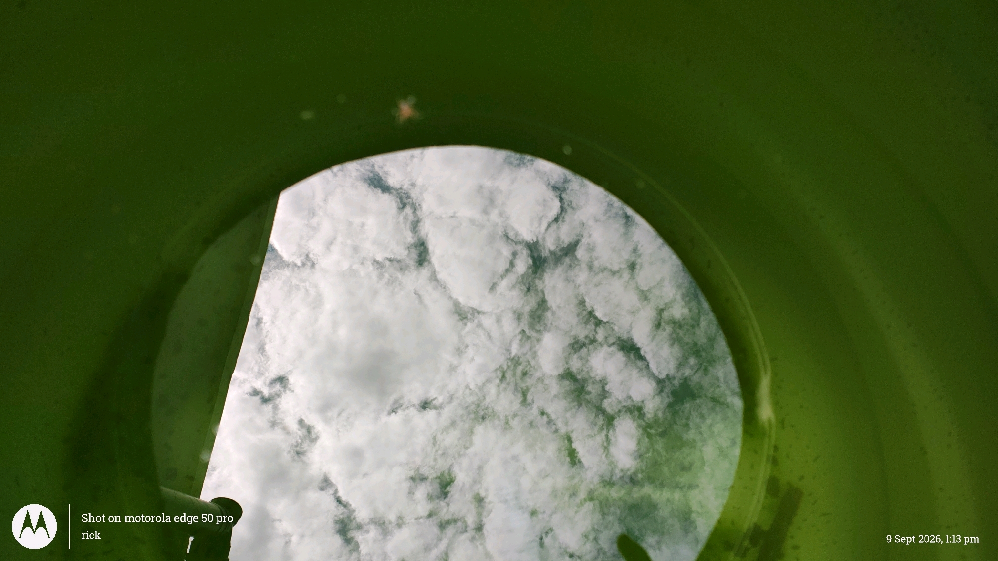 The image is a low-angle shot looking up at a cloudy sky through a curved, green structure. The green structure takes up the majority of the frame, forming a tunnel or opening. White and gray clouds fill the circular opening in the structure. In the bottom left corner, there is a Motorola logo and text that reads "Shot on motorola edge 50 pro rick." In the bottom right corner, there is a timestamp reading "9 Sept 2026, 1:13 pm." The image appears to be taken outdoors in Ishyrwat, India.
