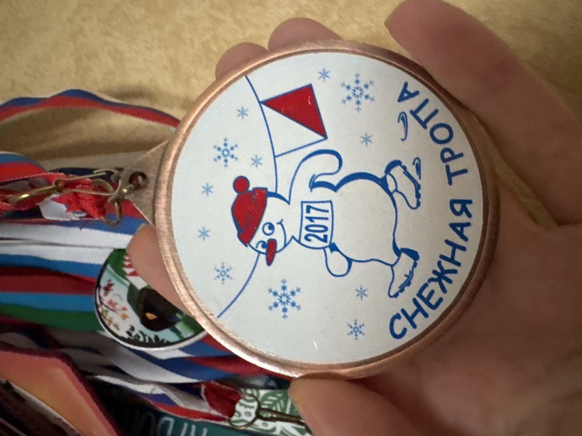 A close-up shot shows a person's hand holding a medal with a snowman design. The medal is circular with a copper-colored rim and a white center featuring a blue outline of a snowman. The snowman wears a red hat and scarf, has a carrot nose, and is depicted on skates. It holds a string with a red triangular flag. The year "2017" is displayed on a white tag around the snowman's neck. The text "СНЕЖНАЯ ТРОППА" (Snowy Trail) is written in blue Cyrillic letters around the bottom half of the medal. In the background, a collection of other medals with colorful ribbons is visible, suggesting participation in an event or competition. The background appears to be a textured, light-colored surface, possibly a wall or fabric. There is no discernible information in the image relating to the city of Sinyavino, Russia.