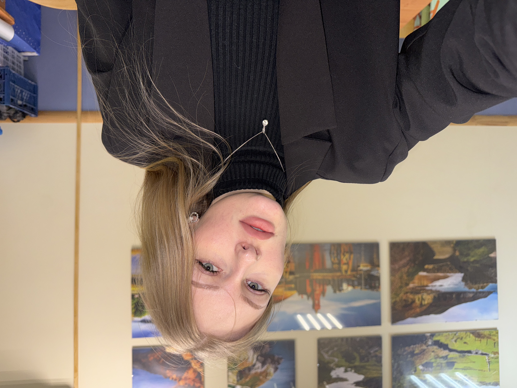 A portrait-style photograph, taken indoors, features a young woman with light blonde hair and light-colored eyes, facing the camera directly. She is wearing a black ribbed turtleneck sweater layered under a black blazer. A delicate silver chain necklace with a small, clear gemstone pendant rests on her sweater. She also wears a small, circular, silver-toned earring with embedded stones in her right ear. Her lips are adorned with a soft pink or peach lipstick, and she maintains a neutral, calm expression.
The background reveals a light-colored wall (beige or cream) decorated with several framed landscape photographs, potentially depicting natural scenery such as rivers, cliffs, and fields, as well as what appears to be a cityscape. In the upper left background, some blue plastic crates or storage containers are partially visible, suggesting an office, classroom, or public institutional setting. The lighting appears to be artificial, consistent with an indoor environment during the day. No text is visible in the image.