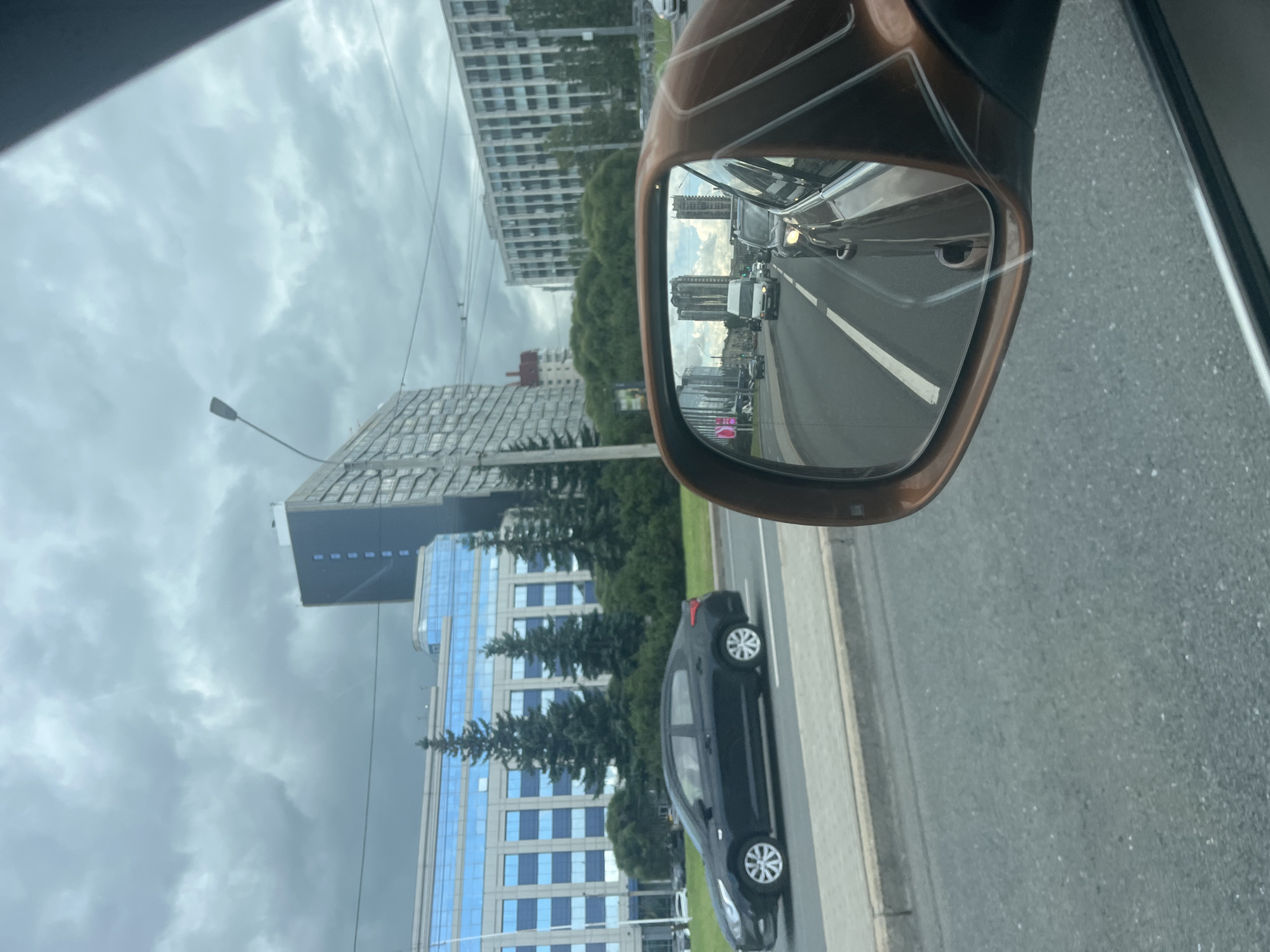 The image captures a view from inside a vehicle on a cloudy day in St. Petersburg, Russia. A side-view mirror reflects traffic on a multi-lane road, with several cars and a white van visible. The buildings in the reflection suggest an urban environment. In the foreground, the side of a dark sedan is visible on the road, moving in the same direction as the observer's vehicle. Beyond the road, a large, modern building with a blue-tinted facade and numerous windows dominates the scene. Tall, dark green coniferous trees are planted in front of this building, and a street lamp stands to the left of it. Further in the background, more buildings of varying architectural styles can be seen, along with overhead wires. The sky is overcast with heavy, grey clouds.