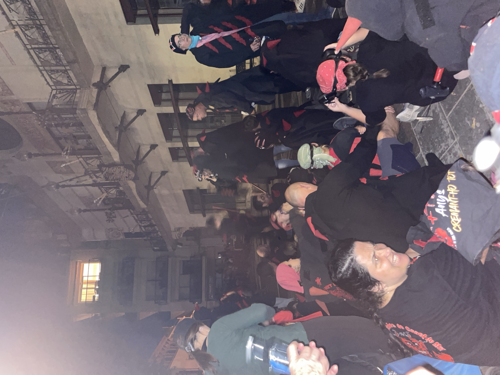 A crowd of people, many wearing black robes and red accents, are gathered on a cobblestone street in front of a building. The building appears to be multi-story with balconies and windows. One window on an upper floor is illuminated. Some individuals are seated, while others are standing. Several people are holding objects, including what appears to be a bottle and a t-shirt. The scene suggests a gathering or event is taking place. The location is identified as Canet de Mar, Spain.