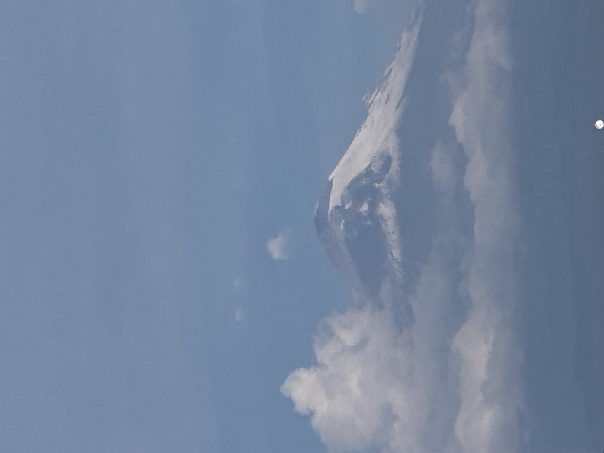 The image shows a mountain partially obscured by clouds against a light blue sky. The mountain appears to have a snow-covered peak. There is a small bright circle in the sky to the right, possibly the moon. Given the prompt specifies a location in San Francisco Ocotlán (Ocotlán), Mexico, the mountain could potentially be near that region, although its specific identity cannot be determined from the image alone.