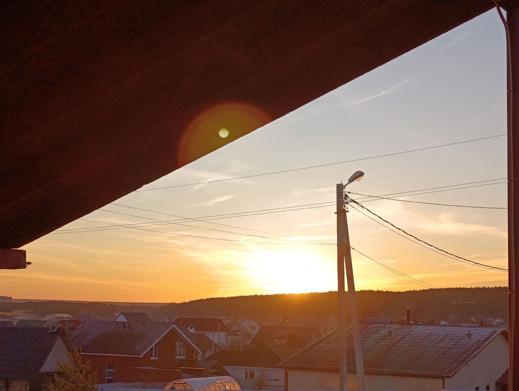 The image shows a sunrise or sunset over the city of Chaikovsky, Russia, viewed from a covered vantage point. A dark brown overhang dominates the upper left portion of the frame. The sky transitions from light blue to a deep orange near the horizon, where the sun is setting or rising behind a tree line. Several rooftops of residential houses are visible below the horizon. A utility pole with electrical wires running across the sky is in the center right of the image.