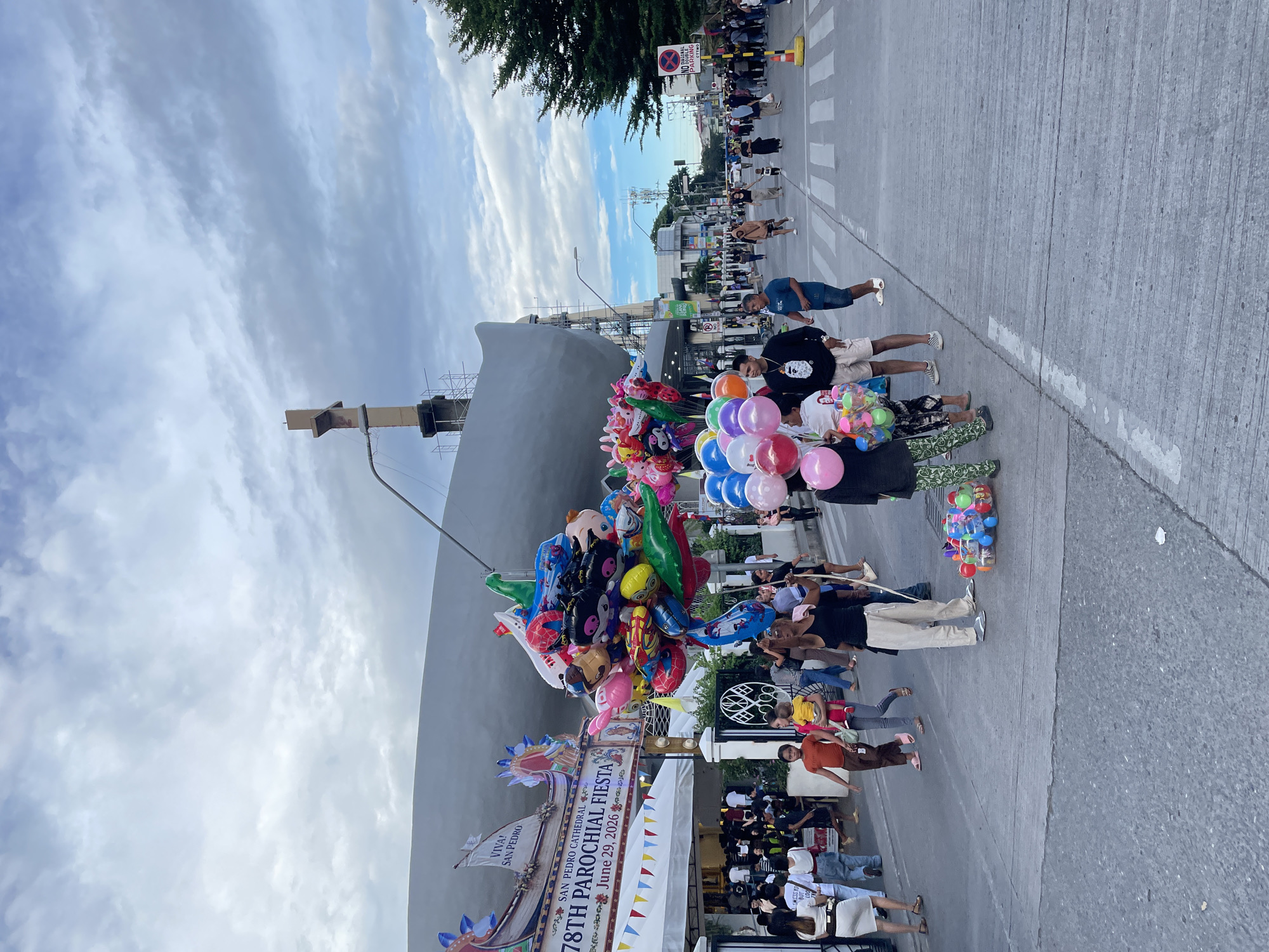 The image shows a street scene at what appears to be a fiesta or festival in Davao City, Philippines. A prominent banner in the background reads "78TH PAROCHIAL FIESTA" and "SAN PEDRO CATHEDRAL," along with "June 29, 2026." A large, modern-looking building is under construction behind the banner, with scaffolding visible.
In the foreground, a vendor is holding a large bunch of colorful balloons, some of which are shaped like cartoon characters. Several people are walking on the street, including families with children. Some are carrying bags or shopping. There are tents set up on the side of the street, suggesting stalls or booths for the event.
The street itself is paved and has pedestrian crossings. Traffic cones are visible further down the street, indicating that the road may be partially or fully closed for the event. The sky is cloudy, with patches of blue sky visible. A large evergreen tree is present on the right side of the image. A "NO PARKING" sign is also visible on the right. The overall atmosphere appears to be festive and crowded.