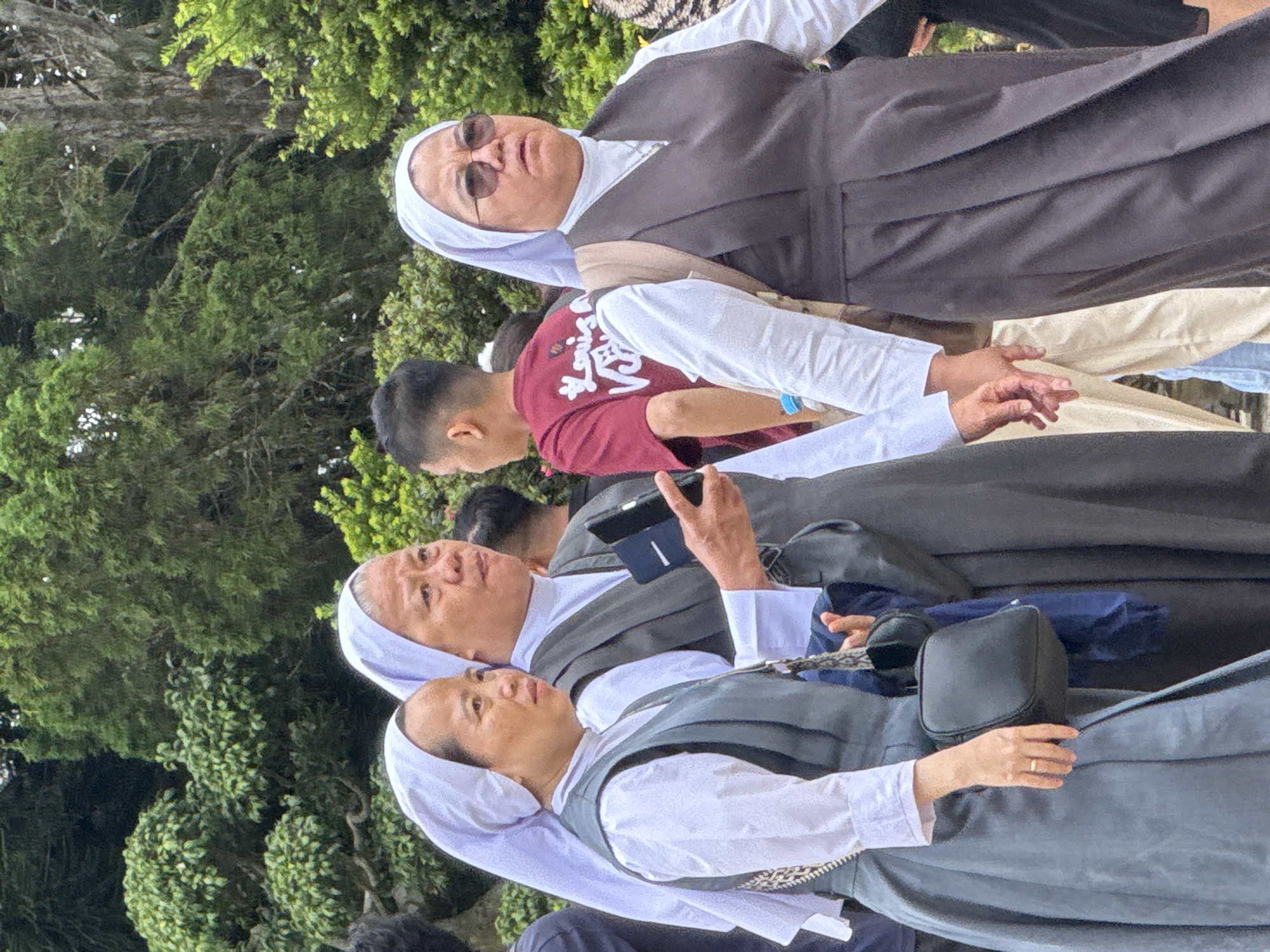 Three Catholic nuns in traditional habits are walking outdoors. The nun on the left is wearing a grey dress and vest over a white blouse, carrying a black purse. The middle nun, also in a grey vest and white blouse, holds a blue tablet. The nun on the right wears a dark grey dress and vest over a white blouse, with sunglasses and a tan backpack. They appear to be walking together, possibly in a public area with trees and other people in the background. This scene could be located in Da Lat, Vietnam, a city known for its numerous religious sites and natural beauty.