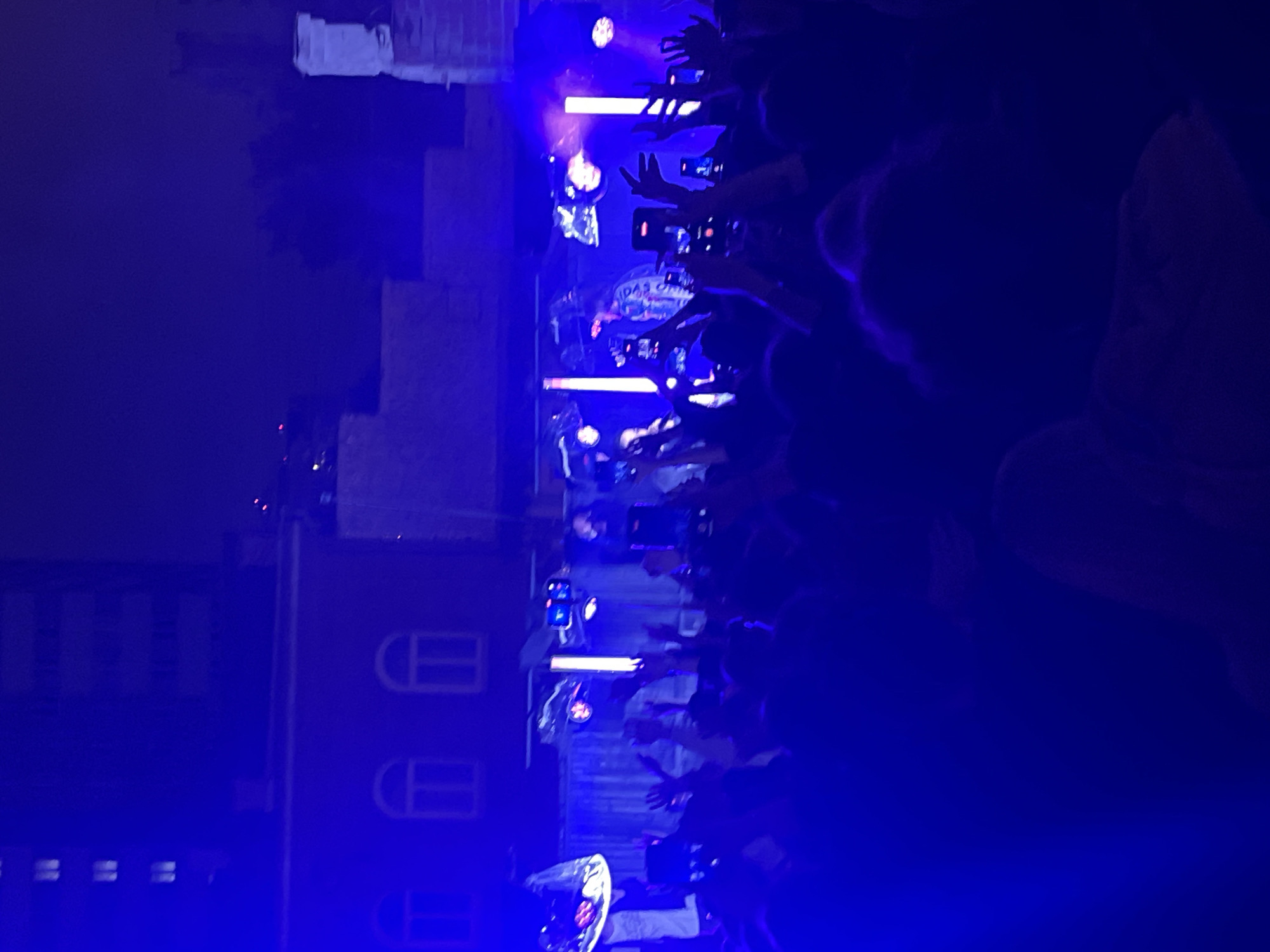 An outdoor concert scene is captured at night, illuminated by vibrant blue stage lighting. A crowd of people stands facing a stage, with many holding up cell phones to record the event. On stage, a performer is visible, wearing a jacket with some graphical designs. The background shows buildings with illuminated windows, suggesting an urban setting. The lighting is intense, casting a deep blue hue over the entire scene and obscuring some finer details due to the low light conditions and overexposure in some areas.