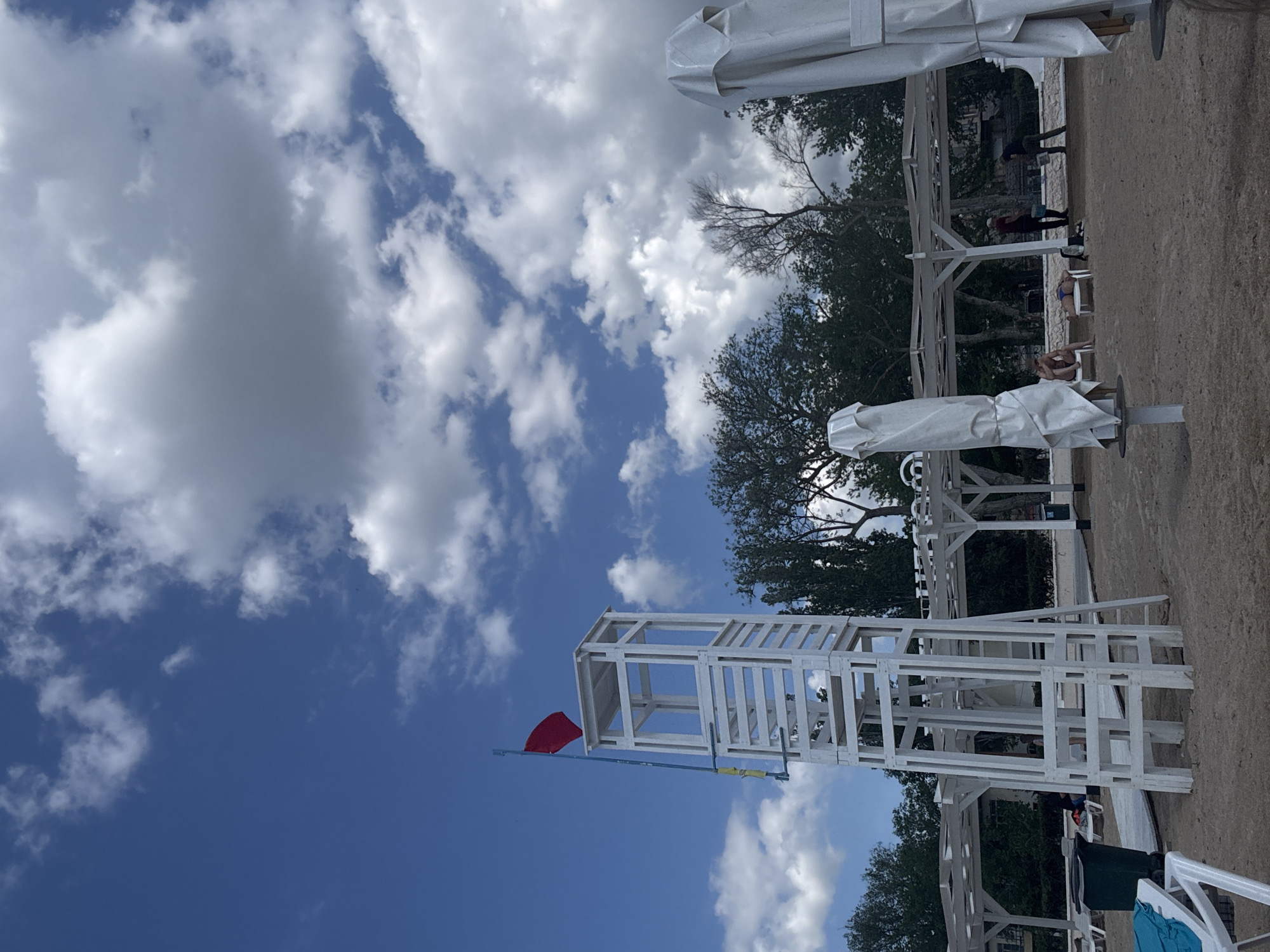 The image depicts a beach scene, likely in Yevpatoriya, Ukraine, given the context. The composition is oriented vertically. A white lifeguard tower with a red flag is visible in the foreground. A blue sky with scattered white clouds fills the upper portion of the frame. In the midground, there are trees and white sunshades. People can be seen lounging on the beach and walking along the shore. The beach appears to be sandy. The scene is outdoors, on a sunny day.