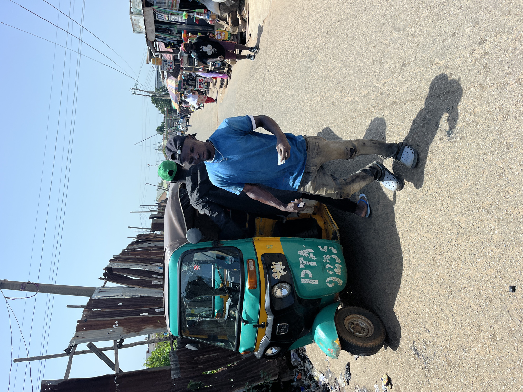 A medium shot captures a street scene on a sunny day. In the foreground, a young man wearing a blue t-shirt, ripped jeans, and grey clogs walks towards the viewer, holding a small white object in his right hand. To his left, a teal and yellow auto-rickshaw is parked, displaying the license plate "PTA-9255" in white lettering. The rickshaw has a brown roof and appears to be slightly damaged, with a cracked windshield. Behind the rickshaw, another person in a black jacket and green cap is visible. The background shows a busy street lined with various stalls and shops, a corrugated metal fence, and power lines overhead. The ground is paved with asphalt, and debris is scattered along the left side of the auto-rickshaw. The overall impression is of a lively, bustling urban environment.