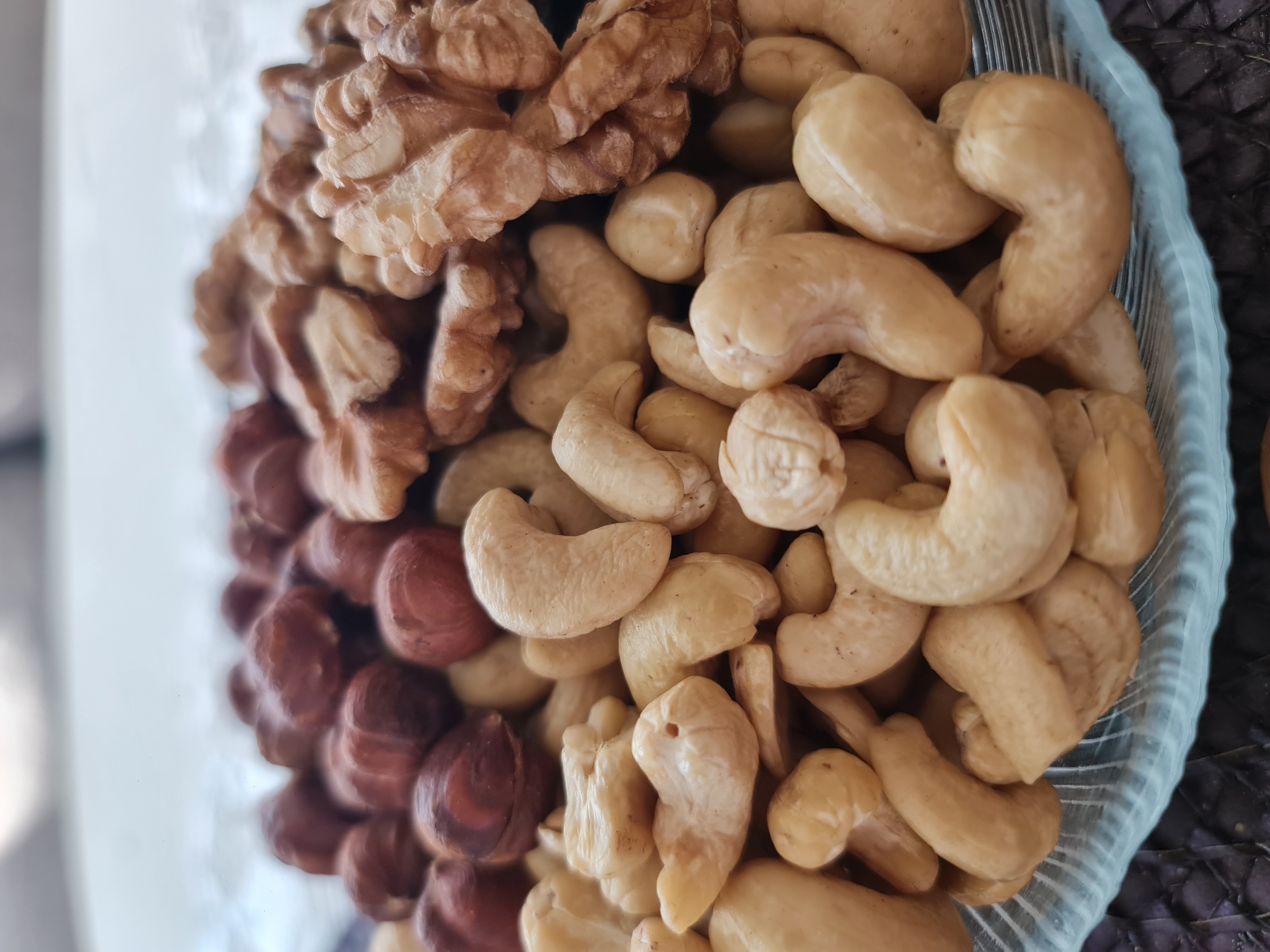 A close-up, eye-level shot shows a variety of nuts piled in a light blue glass bowl. The nuts appear to be hazelnuts, walnuts, and cashews. The hazelnuts are in the upper left corner, dark brown and round. To their right are pieces of walnuts, light brown with their characteristic crinkled texture. The cashews, a pale yellow, fill the majority of the bowl in the foreground, showing their distinctive curved shape. The background is blurred, with hints of a white surface and a dark, textured mat beneath the bowl. There are no people or animals visible in the image. The lighting is natural and appears to be from above. The image is devoid of any contextual information that would indicate its location in Angarsk, Russia, or any other specific city.