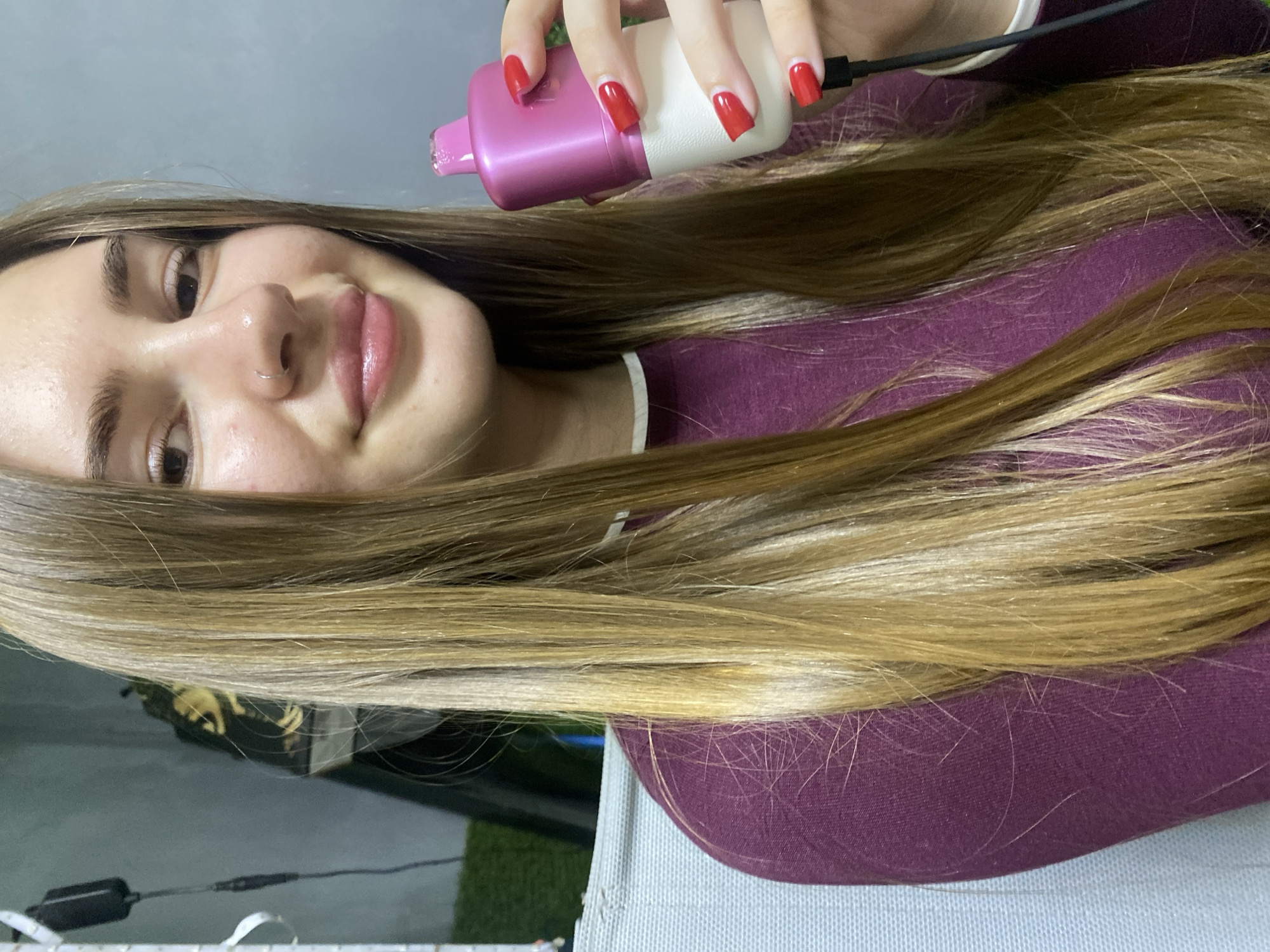 A young woman with long, straight, blonde-brown hair is holding a pink and white device in her right hand. She is wearing a dark purple long-sleeved shirt with a white collar. Her nails are painted bright red. Her left hand is not visible in the frame. The background is blurred but appears to be indoors with a grey wall and some green material. There is no indication of the city of Дьяково, Russia.