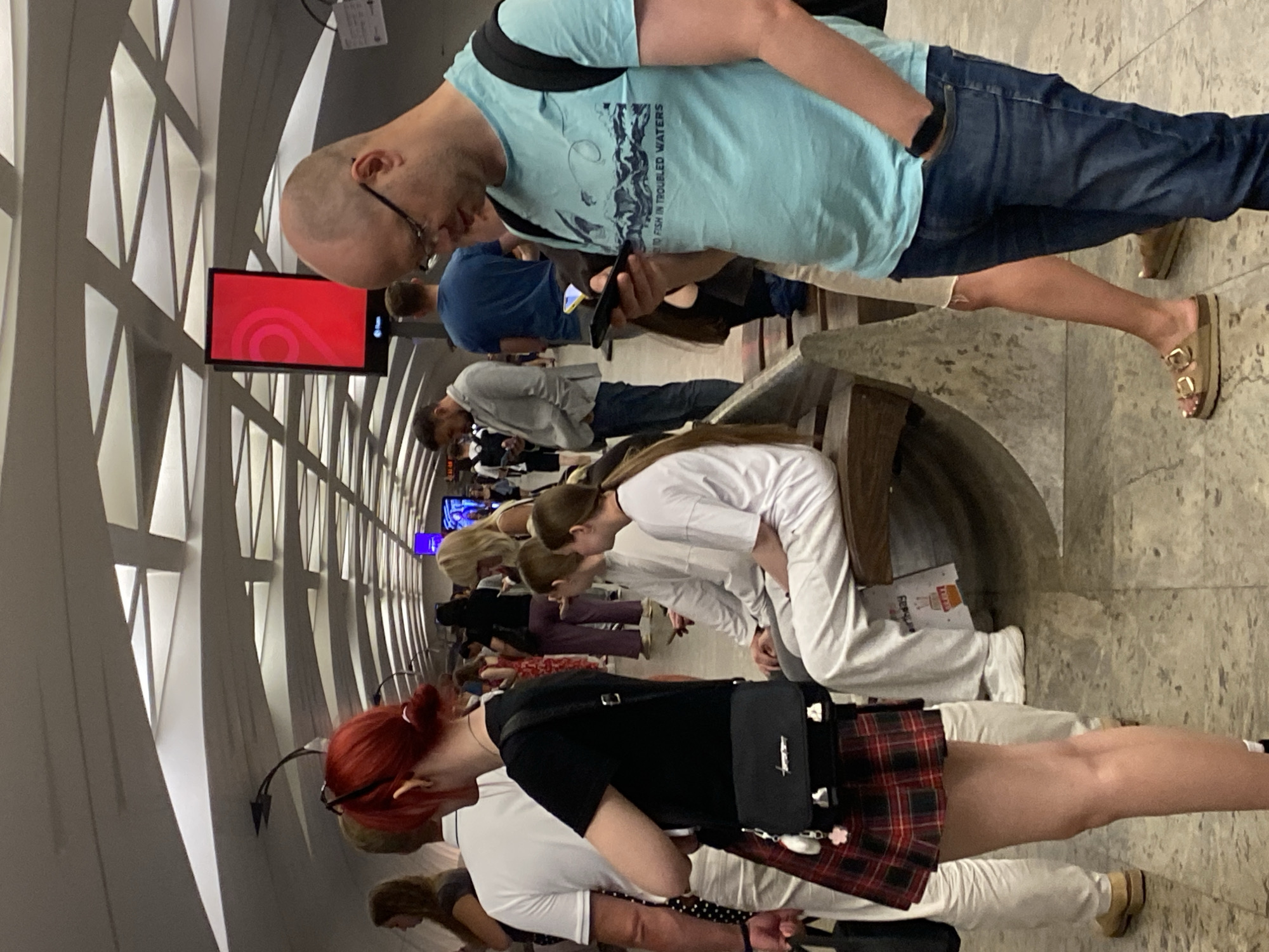 This is a vertically oriented, low-angle photograph taken in Moscow, Russia, depicting a group of people standing and sitting in what appears to be a public transportation station, possibly a metro station, given the architecture with its geometric patterns and tiled flooring. The scene is populated with individuals of varying ages and genders, most of whom are looking down at their mobile phones or engaged in conversation. The overall mood seems casual and perhaps a bit hurried, typical of a transit hub.
In the foreground, a woman with vibrant red hair tied up and wearing glasses is looking down. She has on a black t-shirt, a plaid skirt, and a black crossbody bag. To her left, a person in a white shirt is visible. Behind them, another individual with blonde hair is seen.
Further back, a man in a light blue t-shirt with text and graphics on it is holding a smartphone. He's wearing blue jeans and brown sandals. Next to him, a man in a dark blue shirt and another person in a grey shirt are also present. A red screen displaying a white swirling graphic is mounted on a wall behind the man in the light blue shirt.
The lighting suggests it could be daytime, but the indoor setting makes it difficult to ascertain the exact time of day or weather conditions. The architecture, with its repeating geometric shapes on the walls and ceiling, provides a strong visual element. The ground is tiled with a greyish pattern. The overall impression is of a modern, functional public space filled with people going about their daily routines.