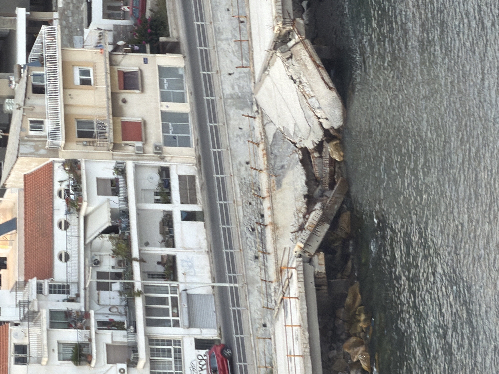 The image depicts the aftermath of a structural collapse at a coastal area in Kavala, Greece. A section of a concrete embankment or promenade has fractured and collapsed into the sea, with large concrete slabs and debris jutting out at precarious angles. The damaged structure appears to be part of a waterfront development or infrastructure.
Behind the damaged embankment, there is a road with a white railing separating it from the collapsed area. A red car is visible on the road, parked near a building. The buildings in the background are multi-story structures with white and cream facades, featuring balconies, windows, and red-tiled roofs. One building on the right appears to be a lighter yellow color. Vegetation, including bushes and some trees, is visible around the buildings and along the coastline. The sea is calm and appears dark gray. The overall scene suggests a recent event causing significant damage to the coastal infrastructure.