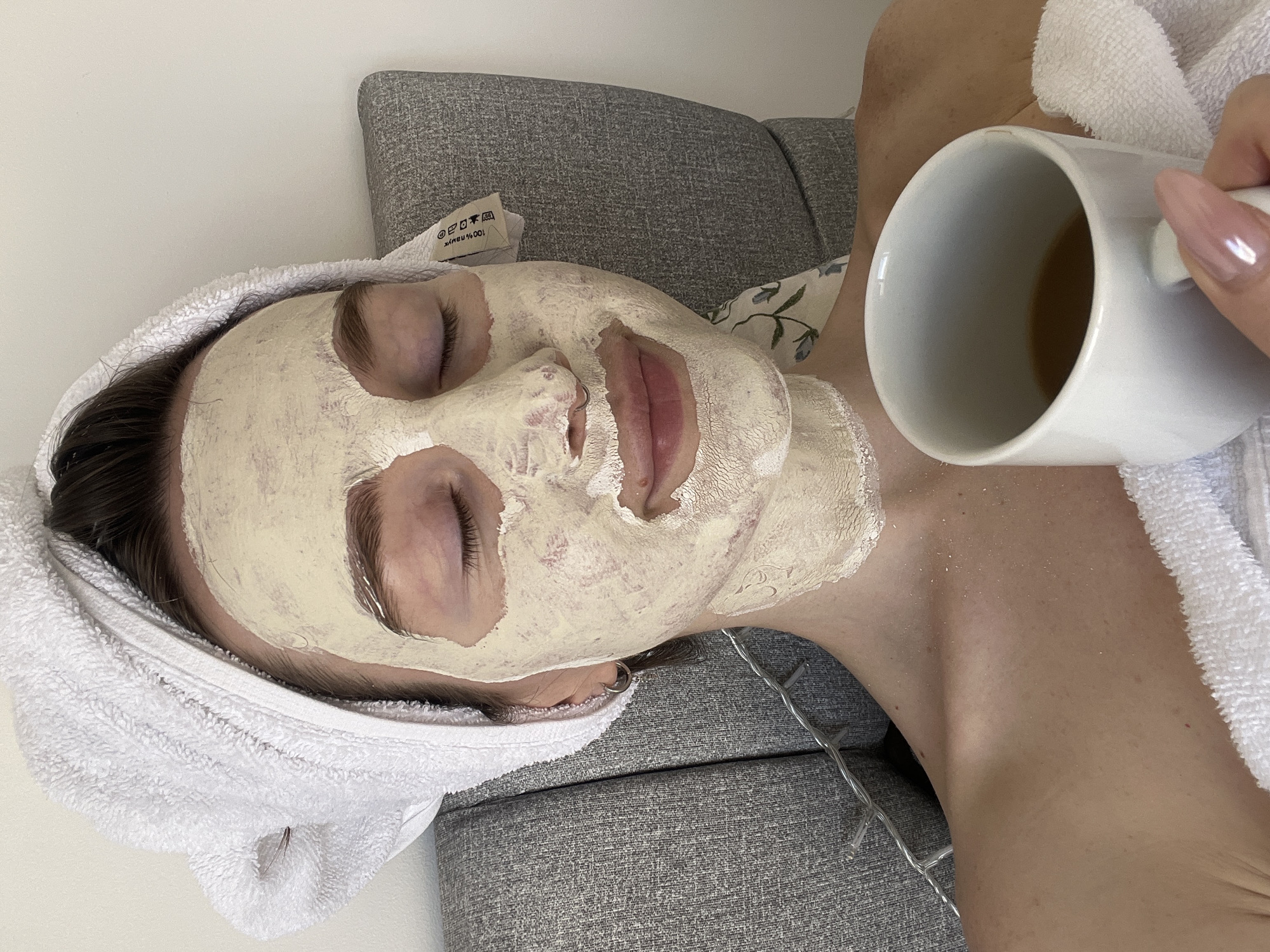 The image captures a close-up, overhead view of a person relaxing indoors. The subject, a woman, is lying on her back, with her head resting on a grey, textured pillow or couch cushion. Her face is covered with a light-colored, possibly clay or cream, facial mask, indicating a self-care or beauty routine. Her eyes are closed, suggesting a state of relaxation.
A white, fluffy towel is wrapped around her head, covering her hair. Her dark hair is visible at the temples and forehead where the mask does not cover. On her left, a hand with long, light pink or nude-colored manicured fingernails holds a white ceramic mug, which contains a brownish liquid, likely tea or coffee.
To the right of her neck, a thin strand of string lights with small bulbs is visible, loosely coiled. The background consists of a plain white wall on the left and the grey textured fabric of the furniture on the right. A small white label with black text and symbols is attached to the grey cushion, positioned near the top of the subject's head. The visible text on the label, which is upside down, includes "100% памук" (Cyrillic for "100% cotton") and standard laundry care symbols (wash, tumble dry, iron, bleach).
The overall impression is one of tranquility and self-pampering. The lighting is soft and even, without harsh shadows.