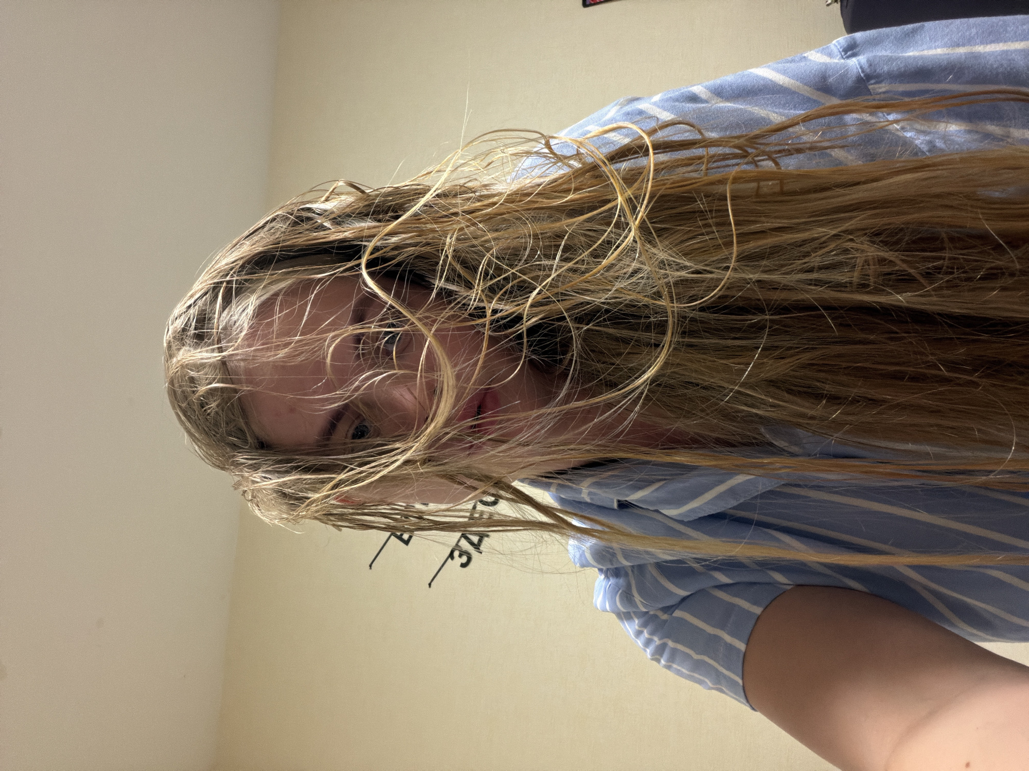 A young woman with long, light brown, wet hair is shown close-up, partially obscuring her face. She wears a light blue and white striped button-down shirt. The background is a plain, light-colored wall. There are some markings on the wall behind her. The location is unknown.