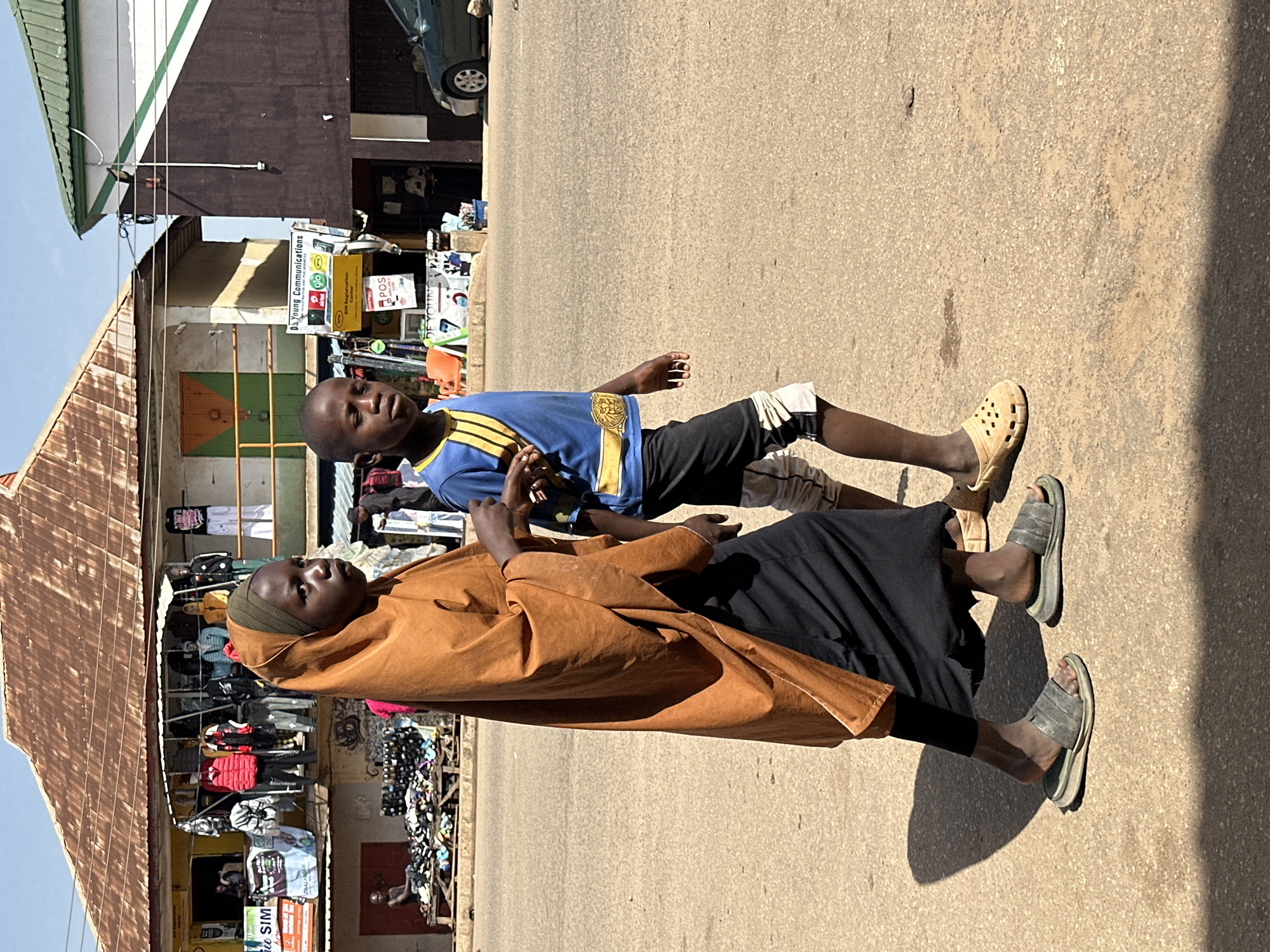 In Bukuru, Nigeria, two young individuals, a female and a male, walk on a dusty, paved street under a bright, clear sky. The female, on the left, is dressed in a long, flowing amber-colored garment that covers her head and body, with a black skirt visible below. She wears dark, worn flip-flops. To her right, the male is clad in a blue t-shirt with yellow accents and black shorts, also wearing tan-colored clogs. They appear to be walking together, with the female slightly ahead and to the left of the male. Behind them, a bustling market scene unfolds. Stalls are set up with racks of clothing and displays of footwear. Buildings with corrugated metal roofs and painted facades line the street. A dark green car is parked further down the road. The overall impression is of a vibrant, everyday street scene in a Nigerian city.