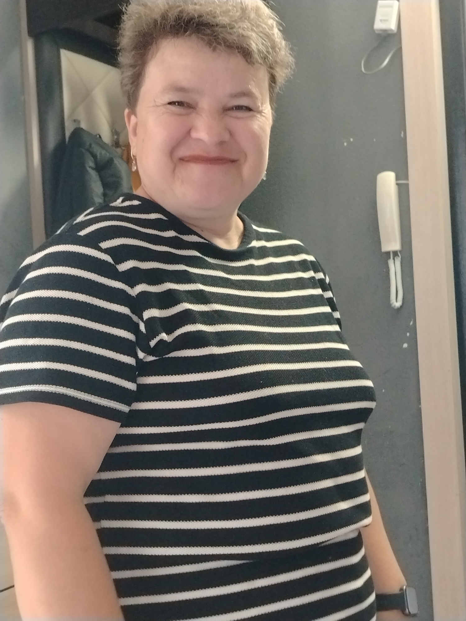An outdoor, medium shot shows a middle-aged woman with short, light brown hair. She is wearing a black and white horizontally striped shirt and a dark smartwatch on her left wrist. The woman is smiling and looking at the camera. She is standing in what appears to be a hallway with gray walls and a light-colored doorframe on the right. A white intercom is mounted on the wall to the right of the woman. A dark jacket is hanging on a hook on the left side of the frame. There are some specks of white paint on the gray wall behind the intercom. The background suggests a typical residential building interior. The image quality is somewhat grainy. The location is unspecified within the city of Noviy, Russia, as there is no definitive visual information to indicate a specific district or landmark within the city.