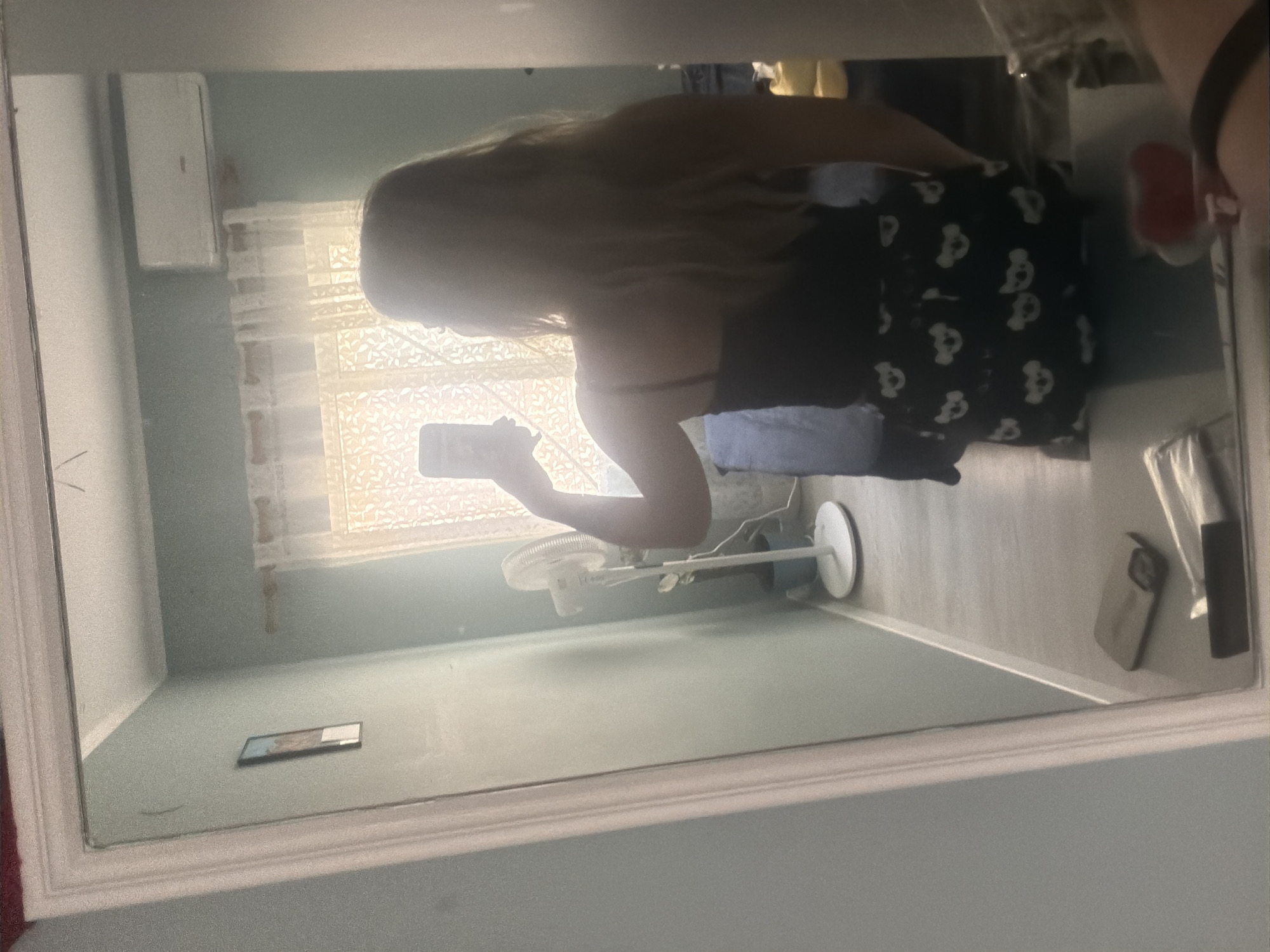 A person is reflected in a full-length mirror, taking a selfie with a phone. The person is facing away from the camera, wearing a black tank top and black shorts with a white koala pattern. The mirror is framed by a white ornate border. The reflection shows a room with light blue walls. To the left, a framed picture hangs on the wall. In the center, a white oscillating fan on a stand is positioned near a window with sheer curtains. Above the window, a white air conditioning unit is mounted on the wall. The floor is light-colored wood laminate. On the floor in the foreground of the reflection, a phone and other objects are visible on the mirror's surface. The scene appears to be an indoor residential setting. This image is unlikely to be from Alicante, Spain, as it lacks any identifying landmarks or architectural features specific to the city.