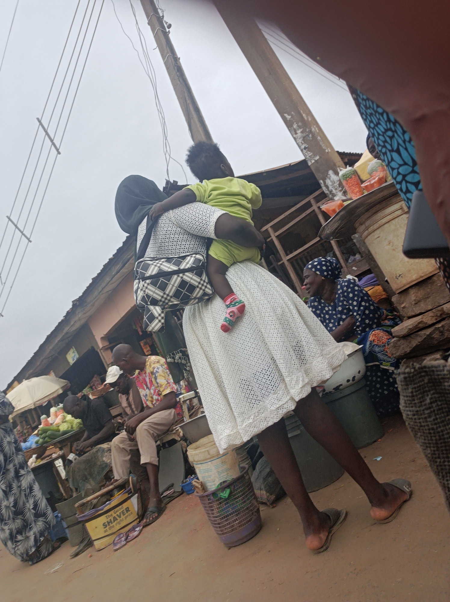 In Narayi, Nigeria, the image depicts a woman in a white dress carrying a child on her back. The child is wearing a light green shirt and patterned socks. The woman has a black and white checkered bag slung over her shoulder and wears a black head covering. She stands in an outdoor market setting with vendors and other people nearby. One vendor sits behind a table displaying vegetables. Banners and signage are visible. Utility poles with wires extend into the sky. The ground appears to be dusty.
