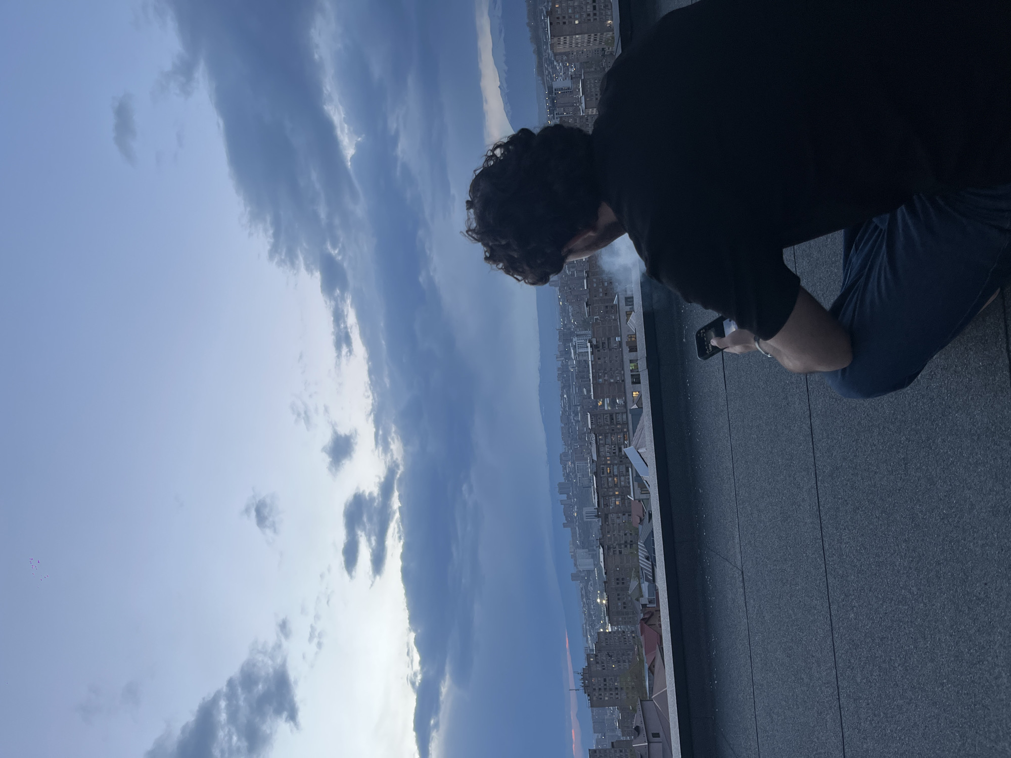 A person is captured from a high vantage point, likely a rooftop, overlooking an expansive urban landscape in Arinj, Armenia. The subject, viewed from behind, has dark, curly hair and is wearing a dark top and blue trousers, kneeling or crouching near the edge of the flat, grey rooftop. They are holding a smartphone in their hands, pointed towards the city below. A faint wisp of smoke or vapor is visible near their face, suggesting they might be vaping or exhaling.
The scene is set under a dramatic, cloudy sky, with shades of blue and grey indicating either late afternoon or early evening. Patches of brighter sky are visible amidst the heavier cloud formations. In the distance, a long stretch of mountains or hills frames the horizon. The city below is densely packed with buildings, many appearing residential or commercial, extending far into the background. Some faint warm light, possibly from setting sun or distant city lights, is visible on the distant horizon above the buildings. The overall impression is one of a vast, tranquil urban panorama, observed by an individual engaged with their phone, perhaps capturing the view or simply taking in the moment. No visible text can be discerned in the image.