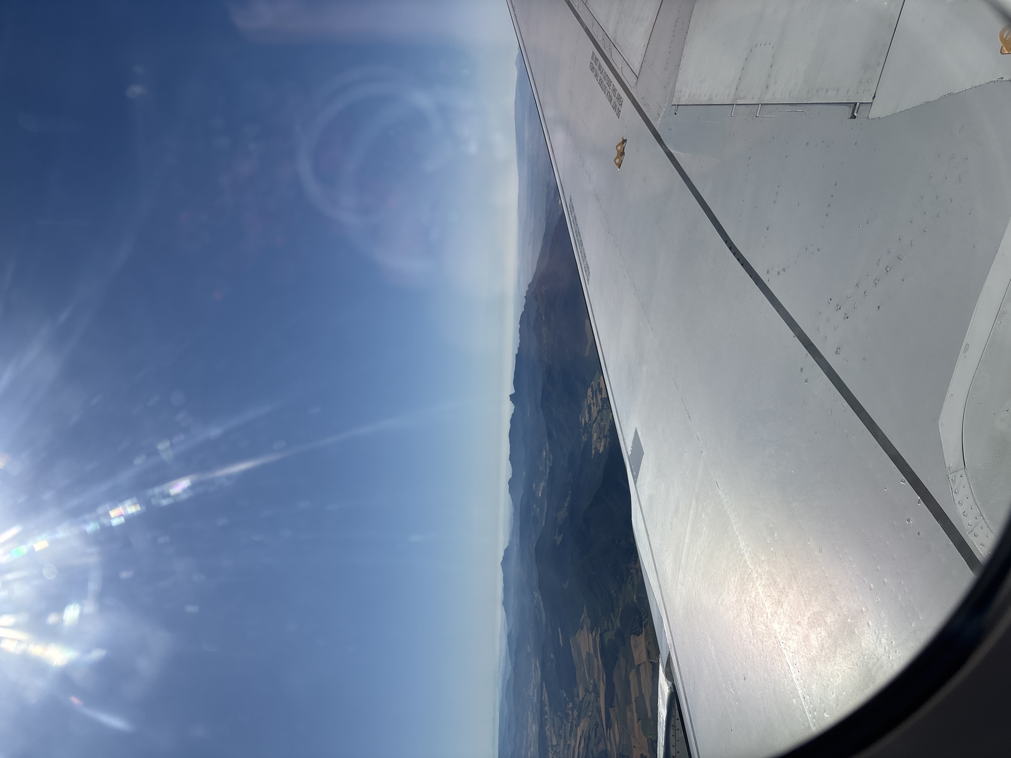 The image is taken from an airplane window, looking out at a landscape of rolling hills and mountains under a clear blue sky. The sun is shining brightly, creating lens flare and reflections on the airplane window and wing. The airplane's wing is visible in the foreground, taking up the right half of the image. The wing is made of white metal and shows rivets and some markings. The landscape below consists of green and brown fields, with dark, forested hills in the distance. The horizon is hazy, with a range of mountains fading into the distance. There are no people visible in the image. The scene depicts a daytime aerial view of a rural landscape. There is no information in the image to suggest the location is the city of Miranda de Ebro, Spain.