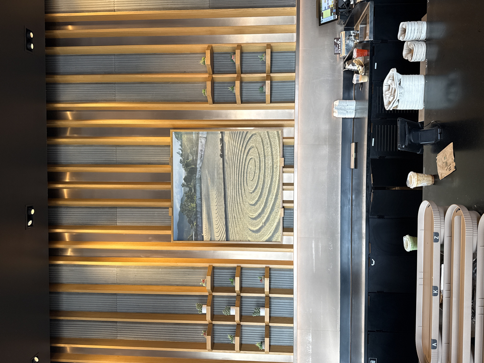 The image captures an interior space in what appears to be a commercial establishment, possibly a coffee shop, situated in Palm Springs, California, USA.
The left side of the frame features a wall adorned with horizontal wooden slats over a textured gray background. A framed image hangs on the wall, portraying an aerial view of a landscape with concentric circle patterns carved into the ground. In addition, a set of small wooden shelves contains miniature potted plants.
To the right, a countertop extends into the space, displaying cups, containers, and other beverage-related items. Behind the counter are black cabinets and equipment indicative of a food and beverage preparation area. On the far right, queue barriers marked with letters "A," "K," and "Z" are visible, suggesting a customer service line. The overall environment seems modern and designed with an aesthetic that combines natural wood elements with sleek, contemporary furnishings.