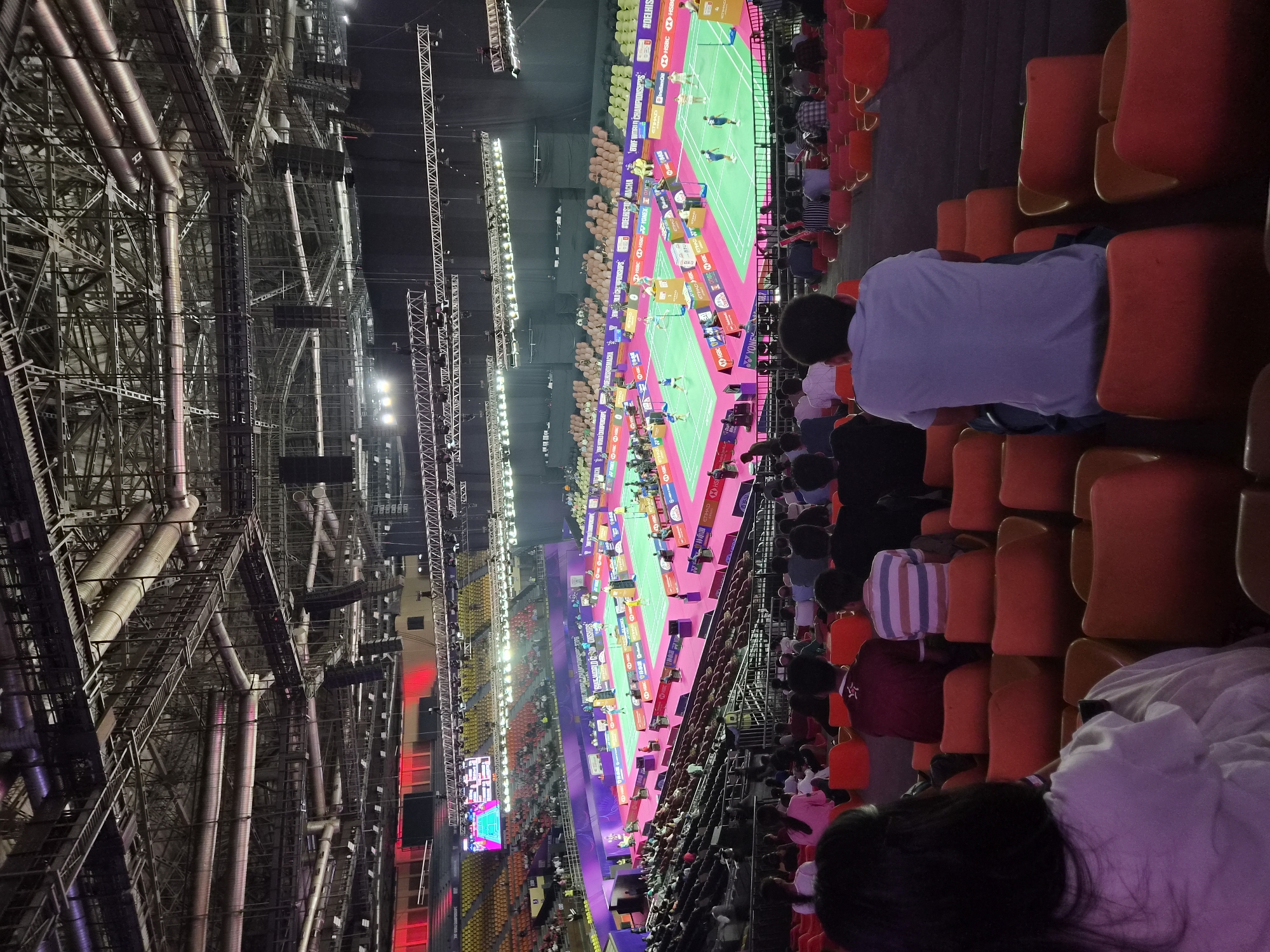 The image depicts an indoor badminton arena during a match, likely the BWF World Championships held in Delhi, India, as indicated by the signage. The arena is filled with spectators seated in orange and red tiered seating. The court below is brightly lit, with multiple badminton courts set up. Players can be seen actively participating in matches on the courts. The ceiling of the arena is a complex structure of metal beams, pipes, and lighting systems. The overall atmosphere suggests a professional sporting event with a live audience.