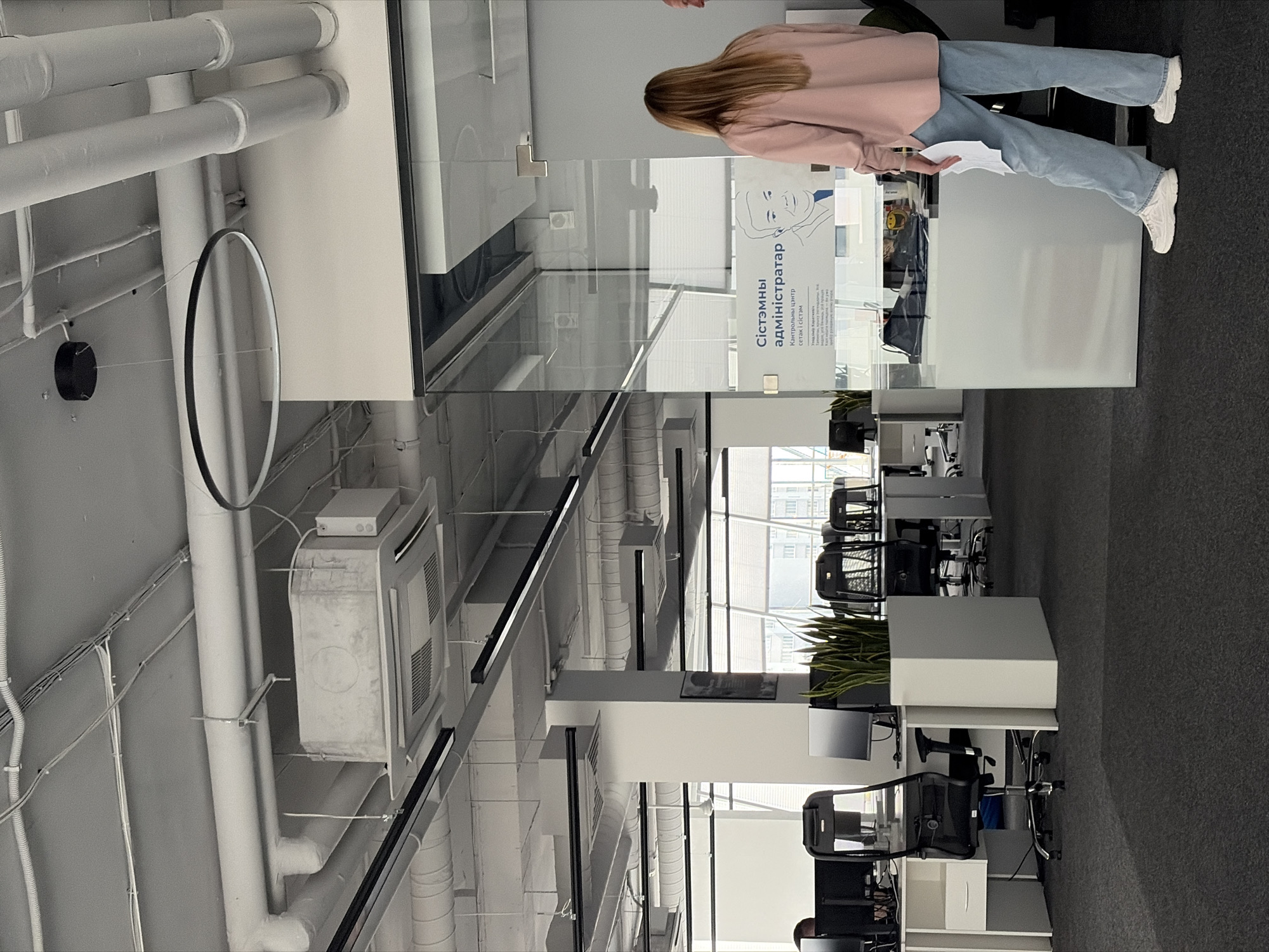 The image depicts an indoor scene of a modern office space. In the foreground, a woman with long blonde hair is standing near a glass partition, wearing a pink shirt and light blue jeans. She is holding a white piece of paper. The office is characterized by exposed ceiling elements, including pipes and ventilation systems, along with recessed lighting. Desks with computer monitors and office chairs are visible in the background, suggesting a workspace. A large plant in a white planter is also present. A sign with text in Ukrainian is affixed to the glass partition. The flooring appears to be dark carpet. The overall impression is that of a functional and contemporary office environment in the city of Бараўлянскі сельскі Савет, Belarus.
