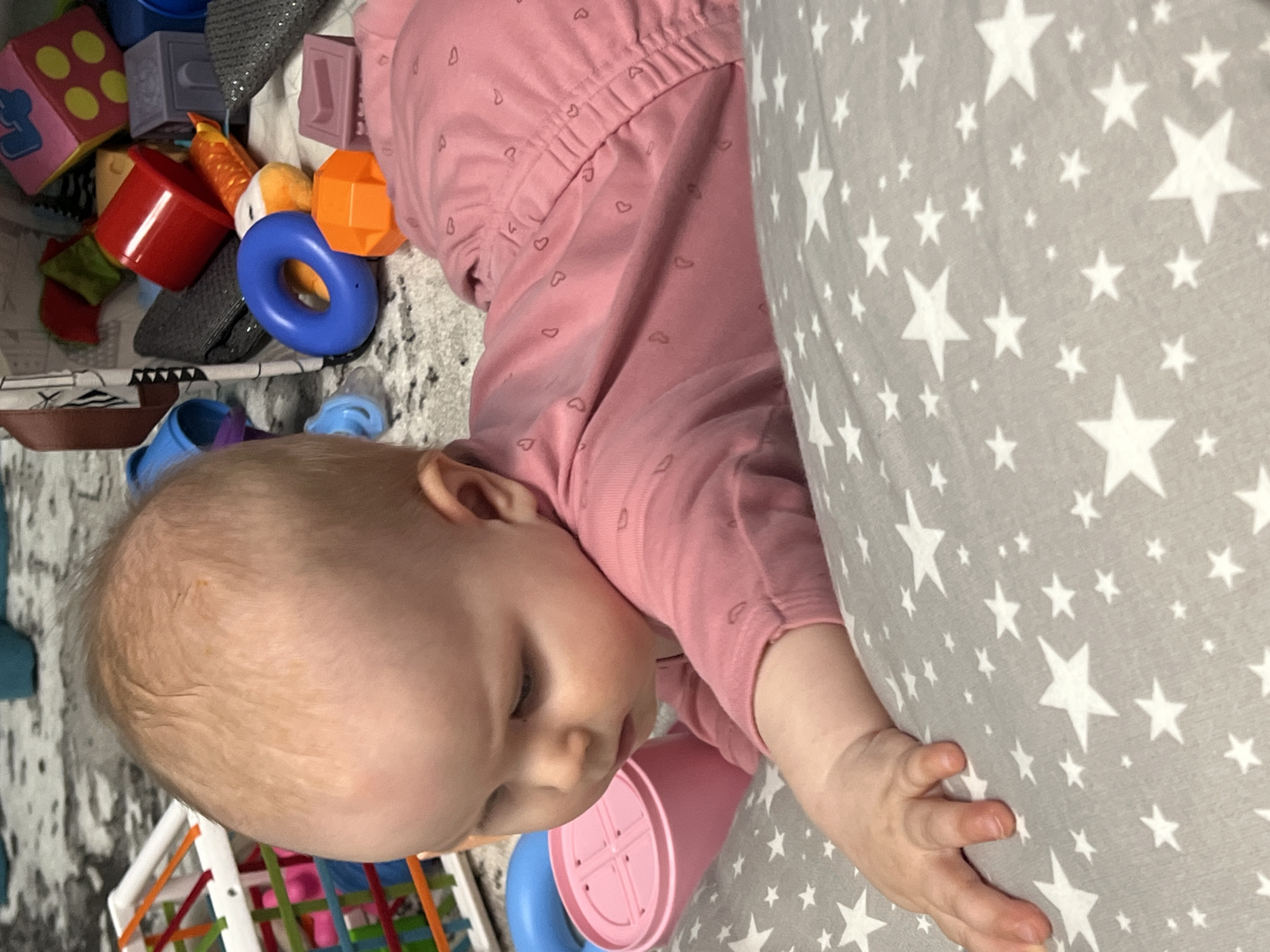 A baby is lying on a gray patterned blanket with white stars. The baby is wearing a pink outfit with heart patterns. The baby is next to a pile of toys, including blocks, rings, and containers, with one container pressed against the baby's cheek. The location cannot be confirmed from this image.