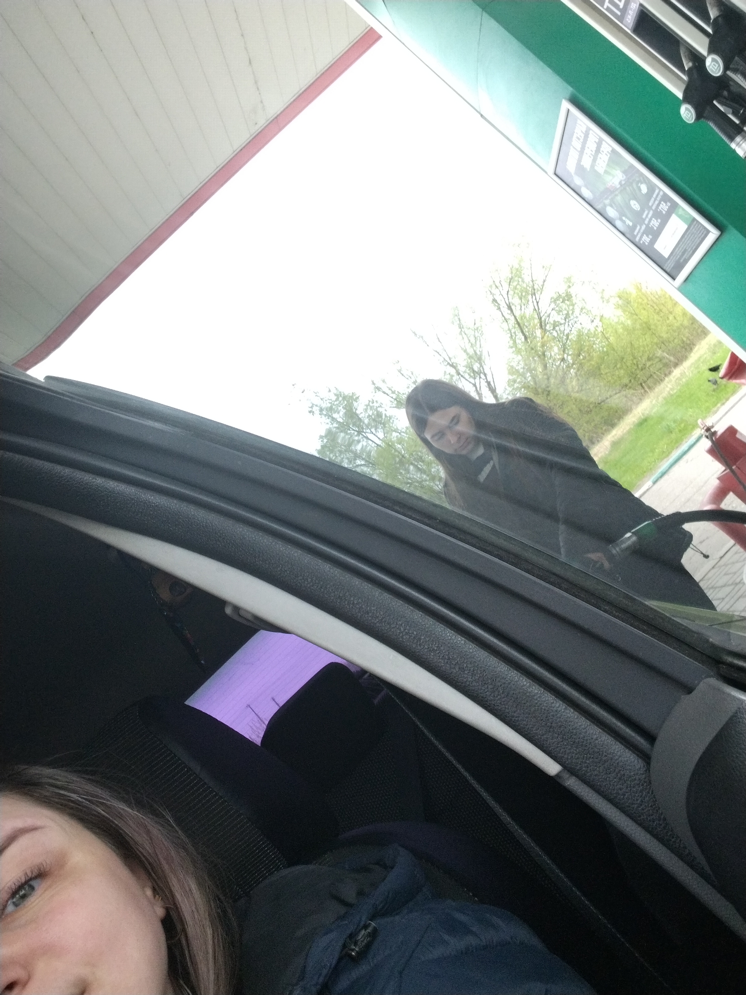 The image depicts two individuals at a gas station, likely in Rusanovo, Russia, though precise confirmation lacks contextual markers. One person is inside a car, visible in the lower part of the frame, with only their face partially visible. The second person stands outside the car, seemingly refilling the car with gas. The gas pump, gas station signage and adjacent greenery are visible behind that person. The image is taken from inside the car.