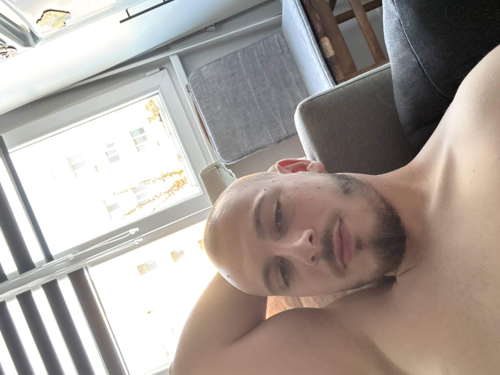 A man with a shaved head and beard is lying on a sofa, with his arm behind his head. He is shirtless. Behind him, a window with blinds is visible, and outside the window, apartment buildings can be seen. A gray upholstered chair is also in the background. The scene appears to be indoors, possibly in an apartment in the city of Stara Zagora, Bulgaria.