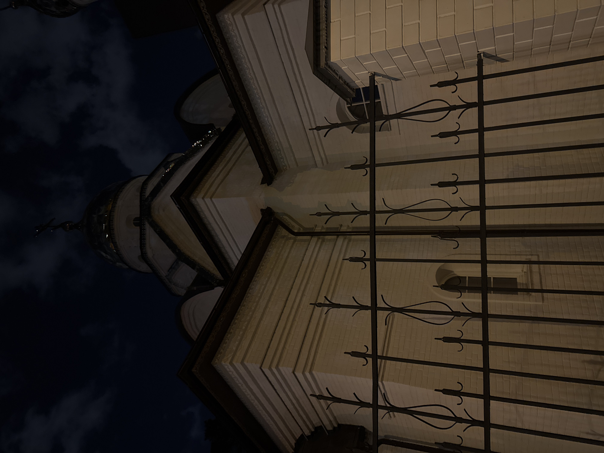 The image shows a low-angle view of a church or cathedral at night, possibly located in Moscow, Russia. The sky is visible with dark clouds. The building is made of light-colored stone or brick, with architectural details such as moldings and cornices. There is a metal fence in the foreground with decorative elements. A dome is visible in the upper left of the image. A window is visible behind the fence.