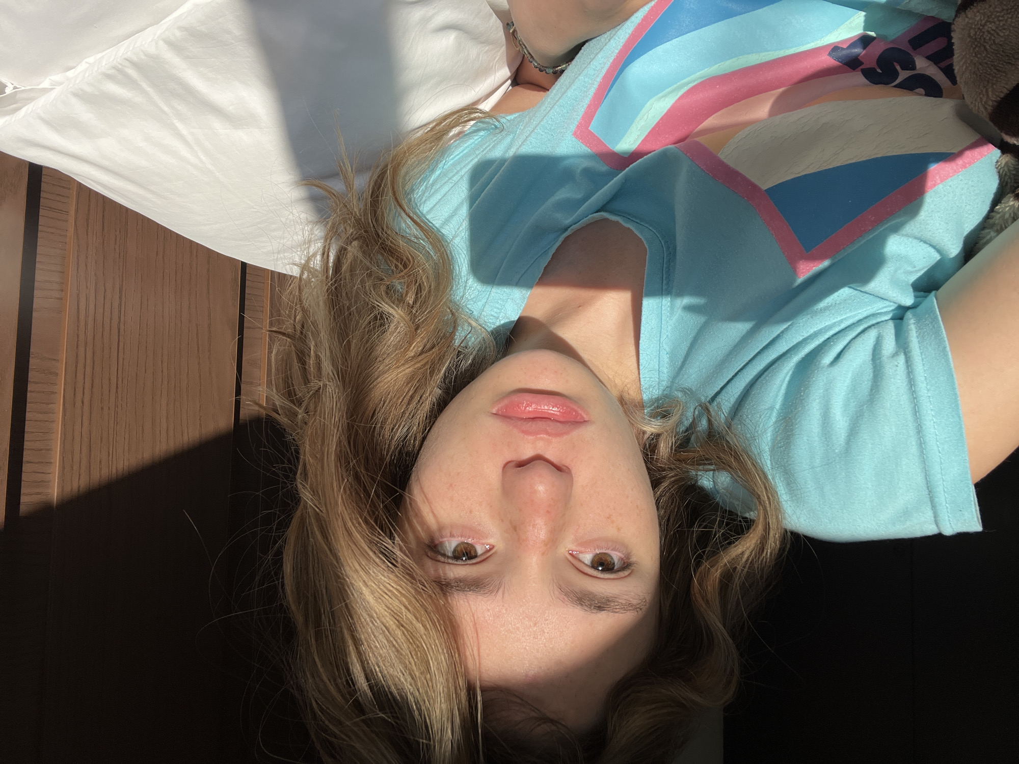 A young woman lies on a bed with a white pillow and a wooden headboard. She is wearing a light blue t-shirt with a colorful design. Part of her face is in shadow. She has light brown hair and brown eyes. The photograph was taken in San Bernardino Tlaxcalancingo, Mexico.