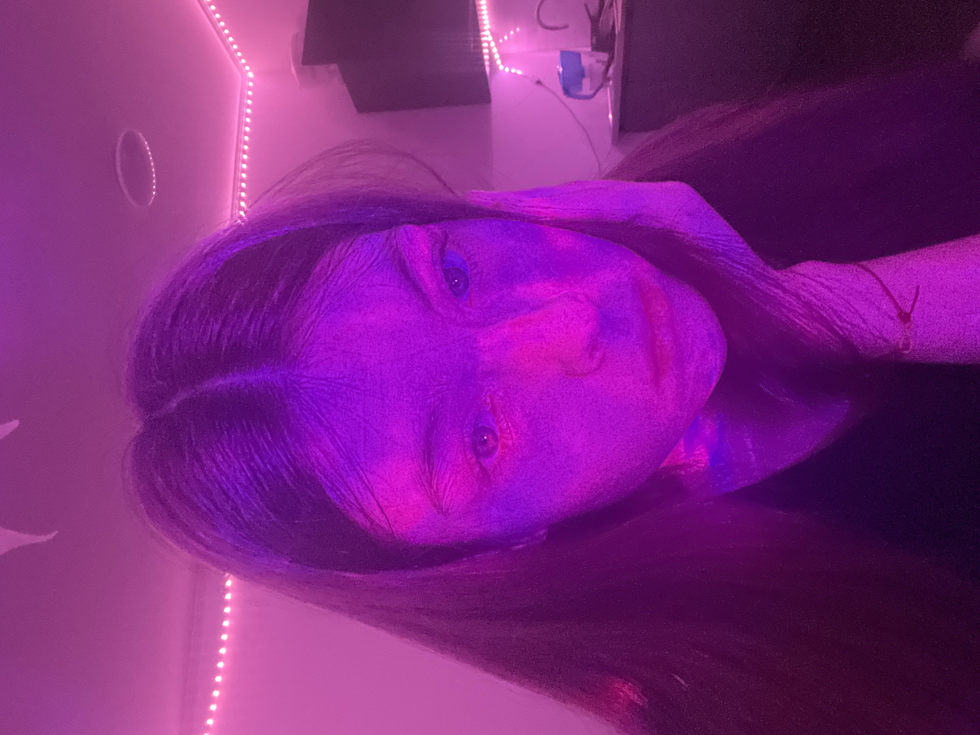 The image portrays an indoor scene dominated by strong pink and purple ambient lighting. A young individual, appearing female, is captured from the chest up, positioned centrally and looking directly at the viewer. She has long, dark hair with a visible part down the middle. Her face is predominantly illuminated by the pink light, with her left hand resting against the left side of her face. A thin, dark band is visible on her left wrist. She is wearing a dark-colored garment.
The background consists of light-colored walls and a ceiling, all heavily tinted pink by the lighting. A strip of small, illuminated pink LED lights is affixed along the upper corner of the wall and ceiling on the right side. On the ceiling, a circular recessed element, potentially a light fixture, is visible. To the right of the individual, a dark, rectangular screen is visible, likely a television or monitor, positioned above a light-colored surface. On this surface, a blue and white mug and several indistinct dark objects are present. The specific location within Saint Petersburg, Russia, cannot be determined from the visual content of this image.