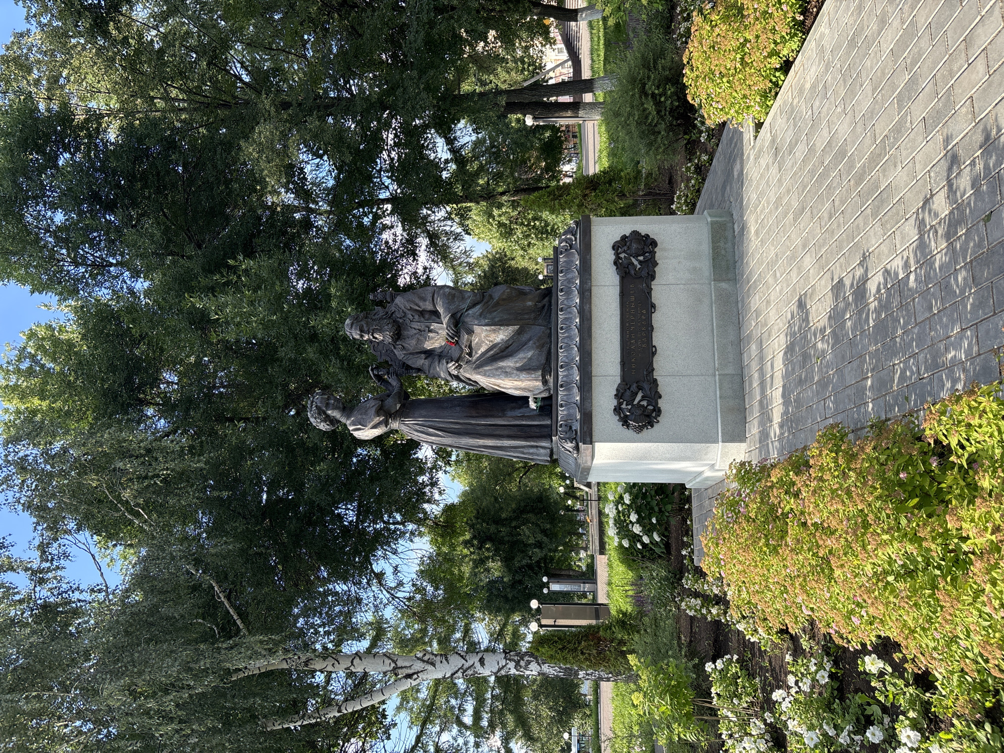 The image shows a statue of Nikolai Chernyshevsky and Varvara. The statue depicts Chernyshevsky sitting on a chair, holding a book and a red flower. Varvara stands next to him, offering him something. The statue is located in a park in Votkinsk, Russia, with lush greenery, trees, and flowers surrounding it. A paved pathway leads to the statue, and there are streetlights and other park amenities in the background. The scene is bathed in sunlight, indicating a clear, bright day.