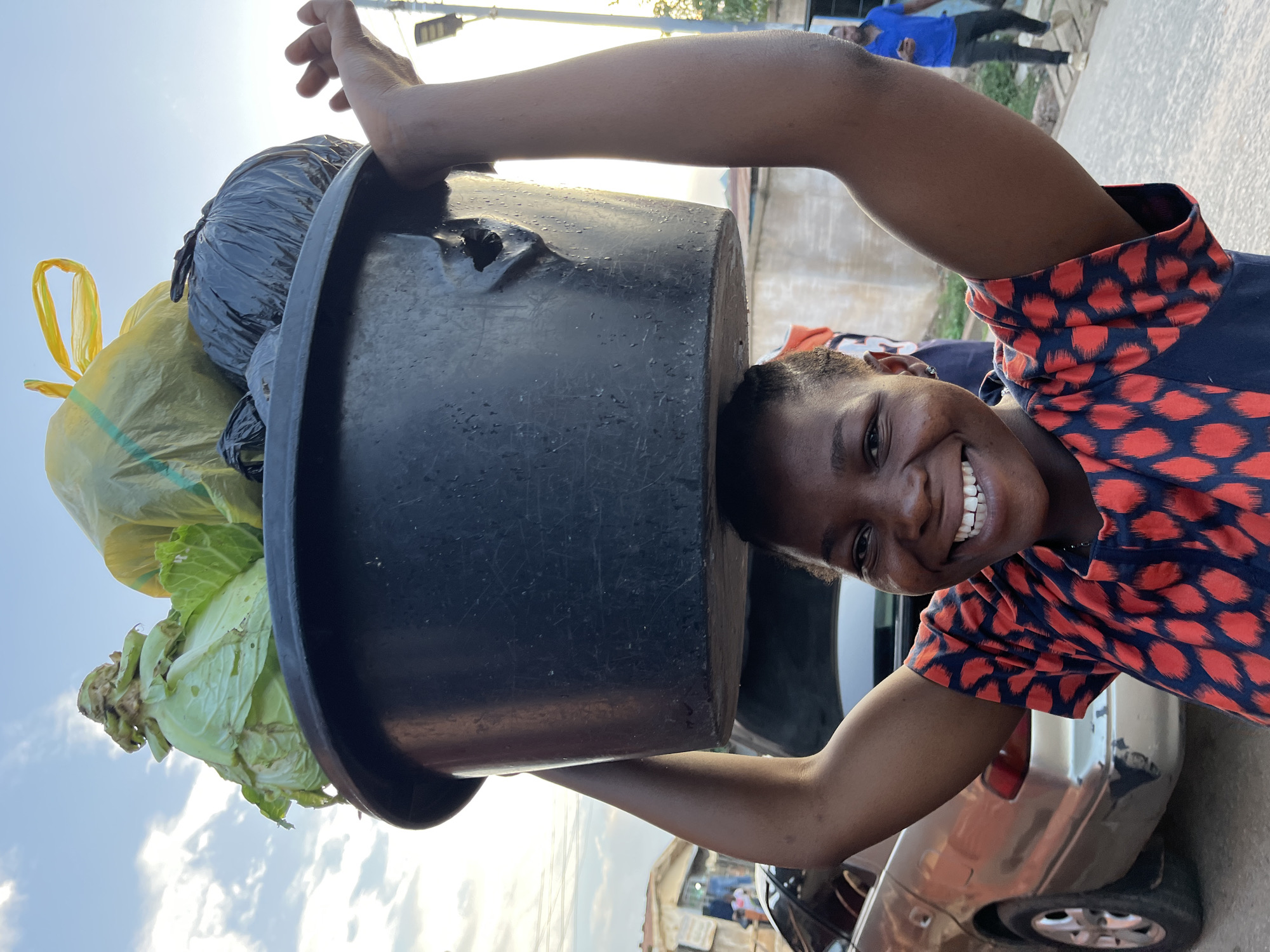 A medium shot, taken in Jos, Nigeria, depicts a smiling young woman with dark skin, dressed in a red and blue patterned dress, holding a large, black, circular container on her head. The container is filled with a mixture of items, including a head of cabbage, yellow and black garbage bags. Her arms are raised to steady the load. The setting appears to be outdoors on a sunny day, with parts of a car, buildings, and a street visible in the background. A person wearing blue is seen walking in the background, along with powerlines visible in the sky.