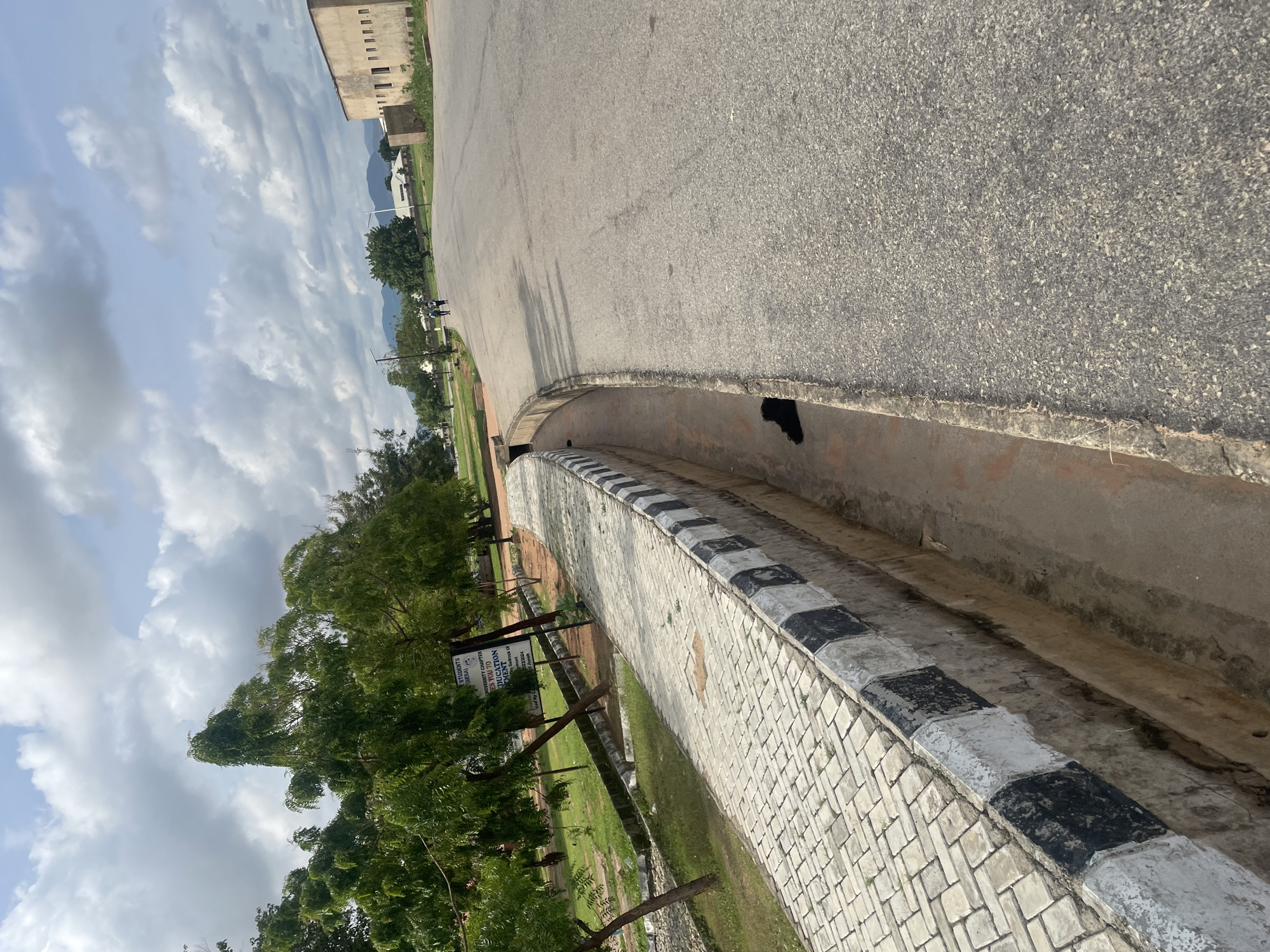 The image shows an asphalt road in Jalingo, Nigeria, with a paved walkway to the left. A drainage channel runs alongside the walkway, bordered by a curb painted with alternating black and white stripes. Green trees line the walkway, and a sign with text is visible behind them. In the distance, buildings and mountains can be seen under a cloudy sky. Two people are walking on the road in the middle distance. A dark object, possibly a piece of litter, is in the drainage channel. The scene appears to be a public area, possibly on a university campus given the sign.