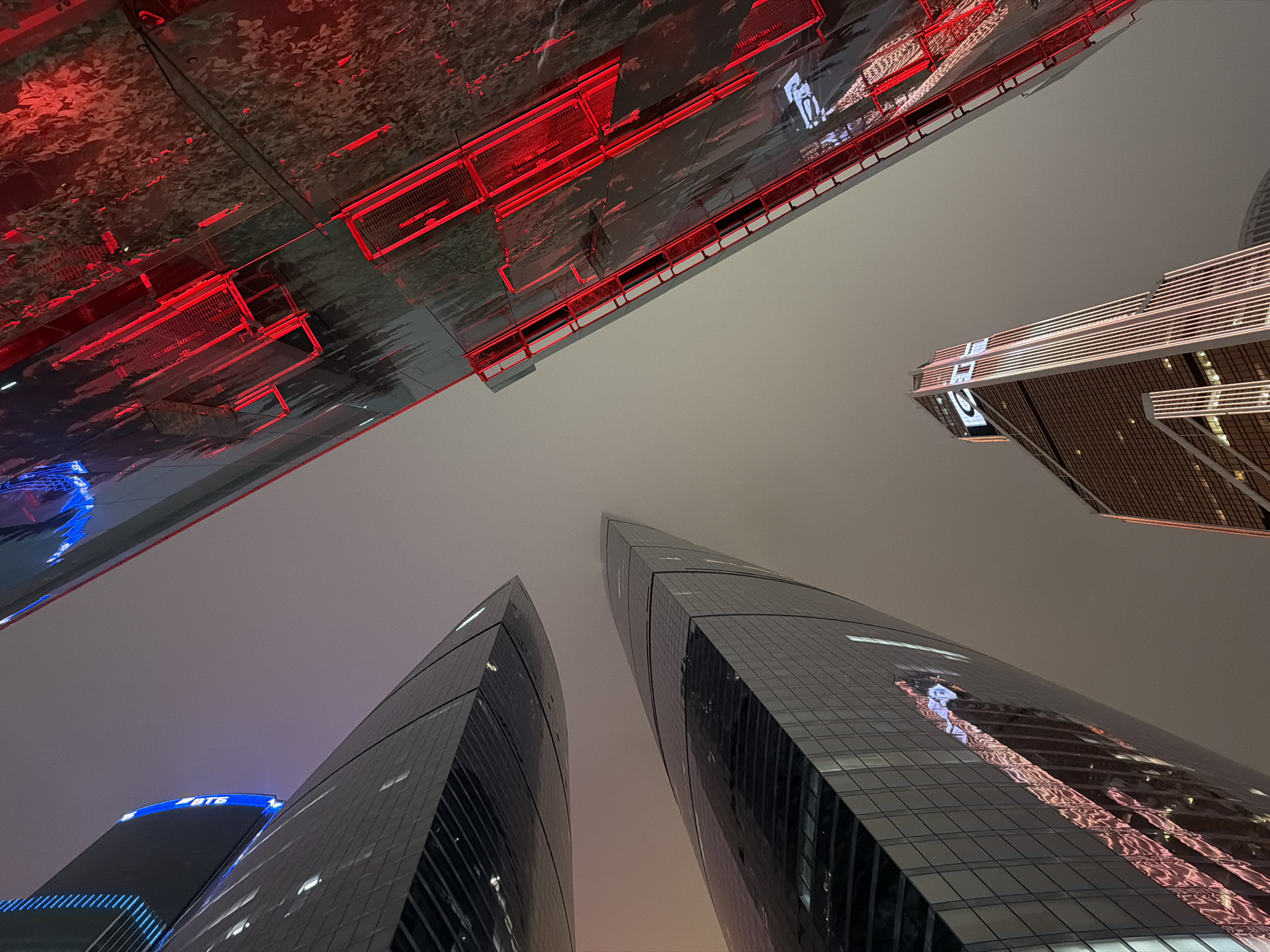 The image shows a low-angle view of several skyscrapers at night in Khimki, Russia. A structure with red lighting and a reflective surface is in the upper left. Two tall, dark glass skyscrapers dominate the lower part of the image. Another lighted skyscraper is in the upper right. The sky is overcast, resulting in a flat grey illumination. "BTB" is visible in blue letters on the top of one of the lower skyscrapers.