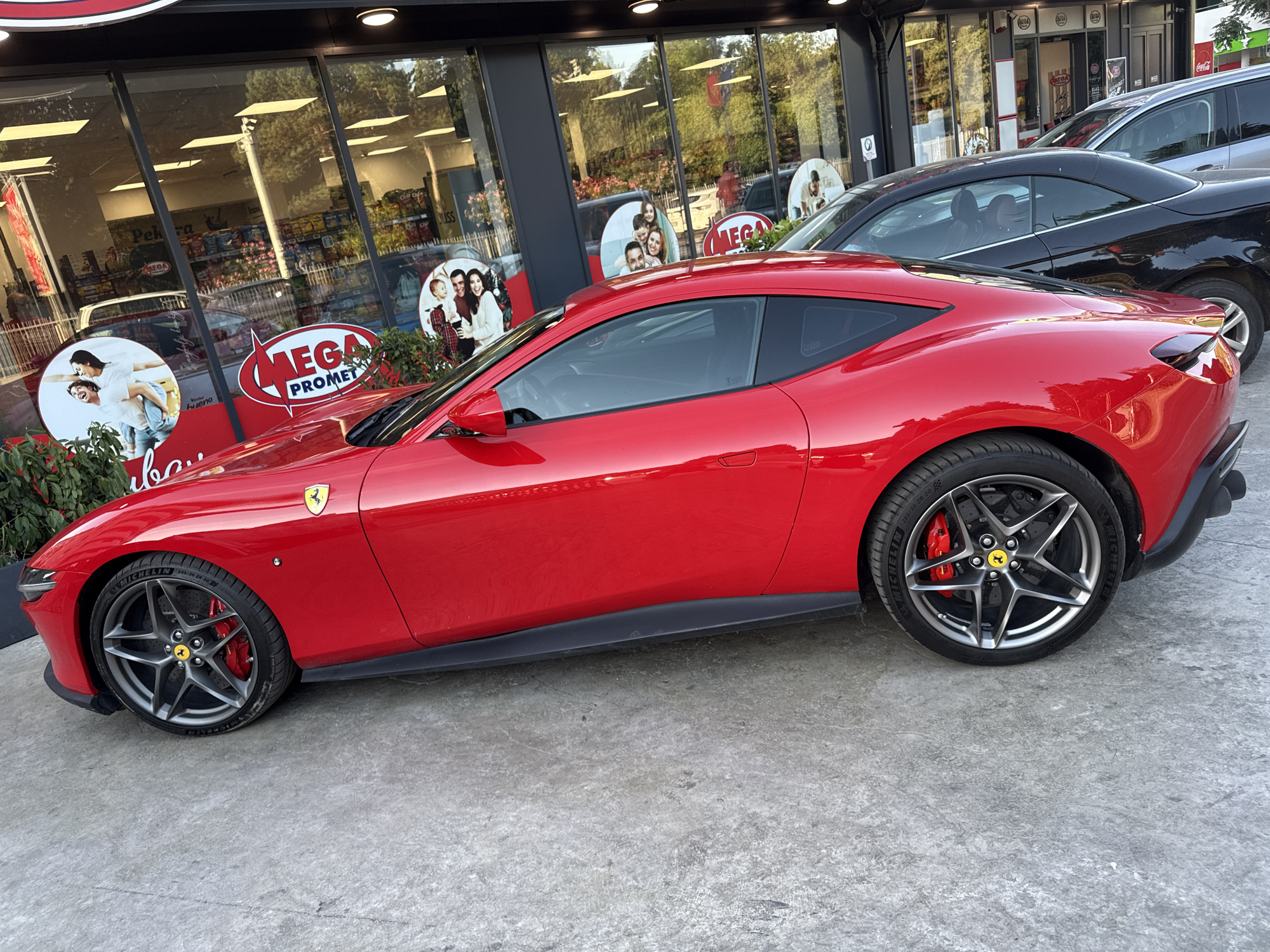 The image captures a vibrant red Ferrari sports car parked in front of a store in what appears to be Budva, Montenegro. The store, identified by "MEGA PROMET" signs in the windows, has a facade with large glass panes reflecting the outside environment. The Ferrari, positioned prominently in the foreground, is parked on a concrete surface. Behind the Ferrari, a dark-colored sedan and a silver SUV are visible, further indicating a parking area. The scene is captured during daylight hours, with natural light illuminating the vehicles and the store. The reflection in the windows showcases the external environment, including some greenery and elements of the town. The overall composition suggests a scene from everyday life in Budva, with a luxury car standing out in front of a local store.