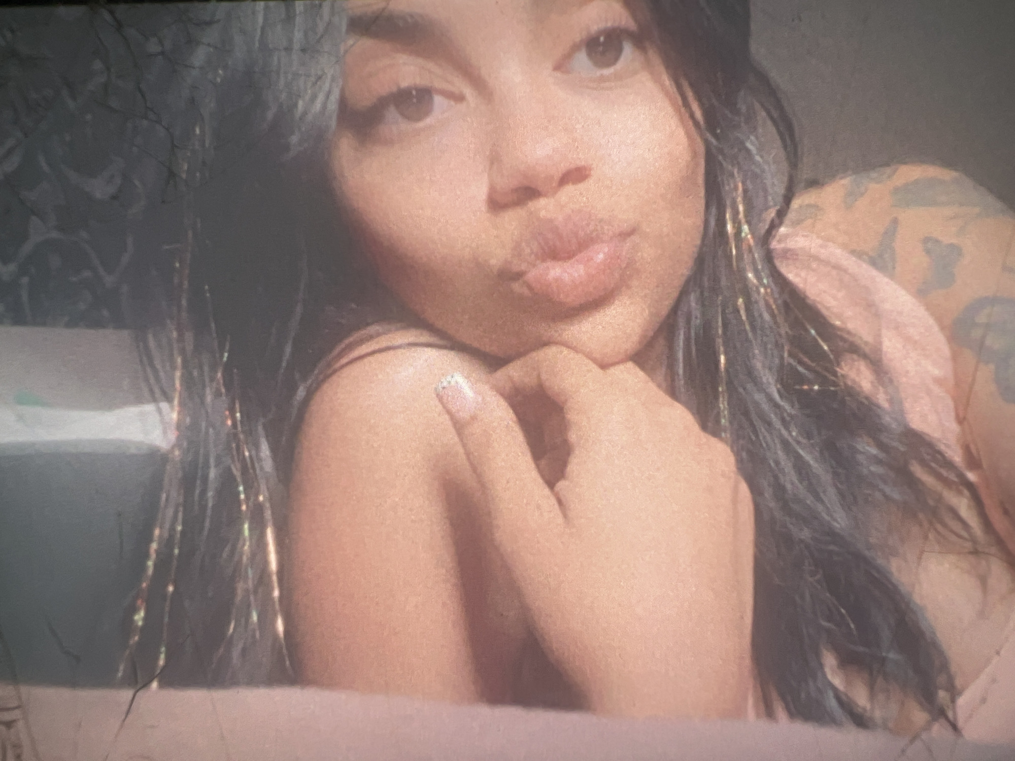 The image is a close-up, vertically oriented photograph of a young woman's face and upper body. She has dark hair, which appears to have some glitter or metallic strands woven into it. Her skin tone is light brown, and she has dark eyes. Her mouth is slightly open, and her left hand is resting on her chin. There is a faint tattoo visible on her shoulder. The lighting is warm and soft, casting a gentle glow on her face. The background is indistinct and appears to be a dark, textured surface. There are some light-colored areas in the background, possibly indicating a wall or fabric. The image appears to be taken indoors, in a casual setting. There is no information in the image to suggest a location in the city of Boone, United States of America.