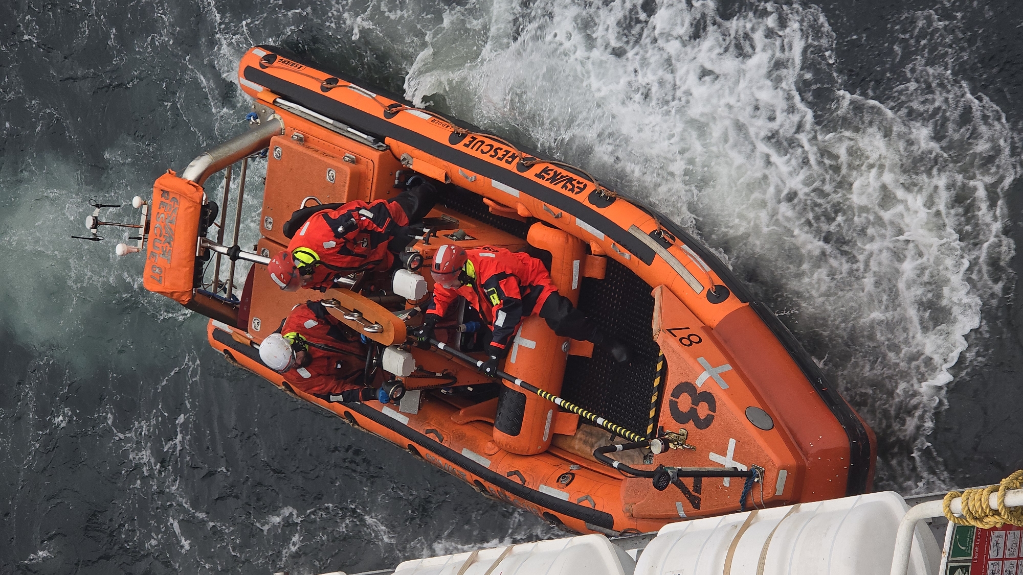 From an elevated angle, the image captures an orange inflatable rescue boat, partly obscured by the frame. Three individuals, clad in red rescue suits and helmets, occupy the boat. One wears a white helmet, another a red one, and the third has a yellow and black helmet. The boat's exterior prominently displays the word "RESCUE" and the number "87." The surrounding environment shows turbulent water, suggesting the boat is in motion, and a portion of a larger vessel is visible in the lower right corner. No city is identifiable in this image.