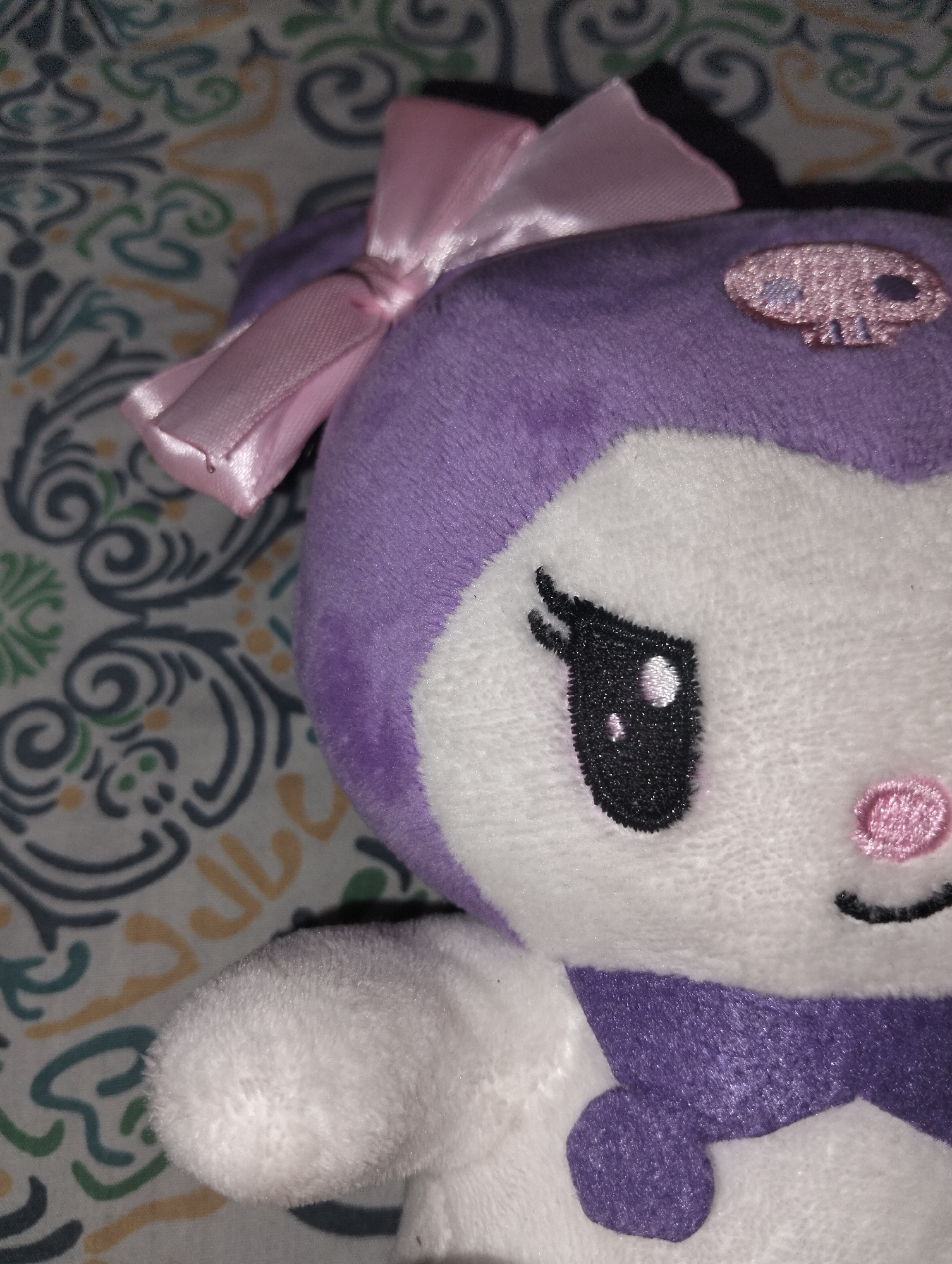 The image displays a close-up of a plush toy, identifiable as the Sanrio character Kuromi, positioned against a patterned fabric background. The toy features a white face, visible right arm, and chest area, contrasted by a purple headpiece and collar. A pink satin bow is affixed to the left side of the toy's headpiece. An embroidered pink skull motif, with black eye sockets and nose/mouth detail, is present on the upper right side of the purple headpiece. The toy's face shows a large black embroidered eye with two distinct white highlights, a small pink embroidered nose, and a thin, curved black embroidered mouth. The background is a light-colored textile, possibly white or cream, adorned with an intricate, repeating pattern of curvilinear and stylized floral motifs in shades of dark gray, teal, and pale yellow/orange. No individuals or active interactions are depicted. The precise location within Dhaka, Bangladesh, cannot be determined from the visual information provided in the image.