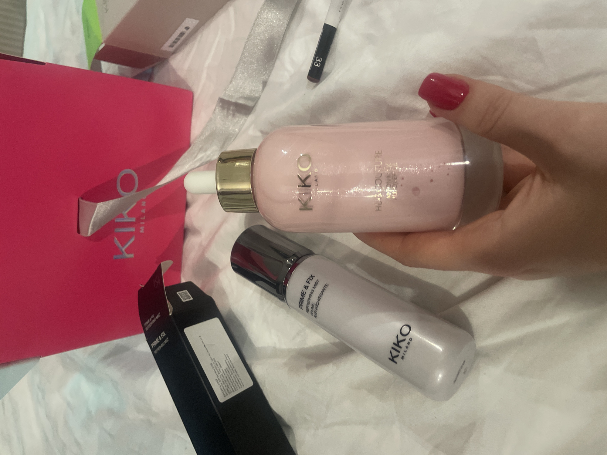 A fair-skinned hand with fuchsia-painted fingernails holds a Kiko Milano Hues Couture liquid product in a pink, clear-bottomed bottle with a gold dropper top. To the left, a Kiko Milano Prime & Fix Refreshing Mist spray bottle is visible. Behind it lies an open black cardboard box for the same Prime & Fix product. In the upper right quadrant, a fuchsia Kiko Milano shopping bag with silver ribbon handles is prominent, displaying the Kiko Milano logo. A white cosmetic pencil with a black cap and the number "33" is situated to the right of the held bottle. All items are arranged on a wrinkled white fabric surface. The scene is an indoor setting within the city of Sofia, Bulgaria.