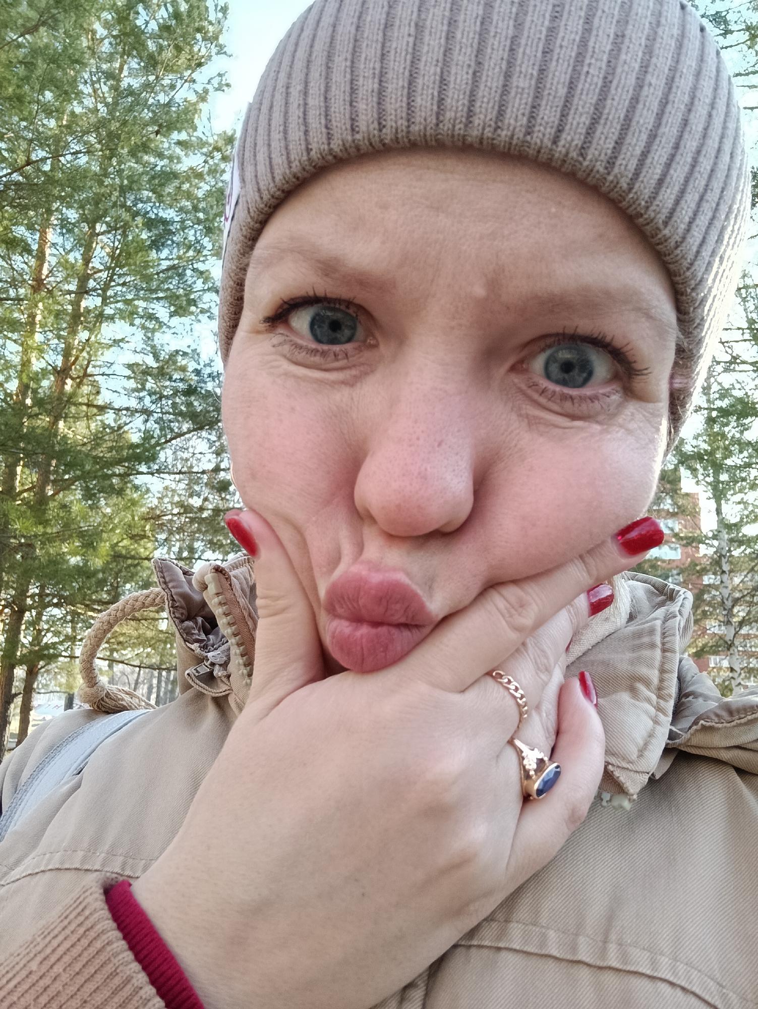 A woman is taking a selfie outdoors, with trees visible in the background. She is wearing a beige beanie, a beige jacket, and a red sweater sleeve is peeking out. She has red nail polish. She is holding her face with her hand, puckering her lips. She is wearing a gold chain ring and a gold ring with a dark stone. Based on visual cues, the location is likely in the city of Chaikovsky, Russia.